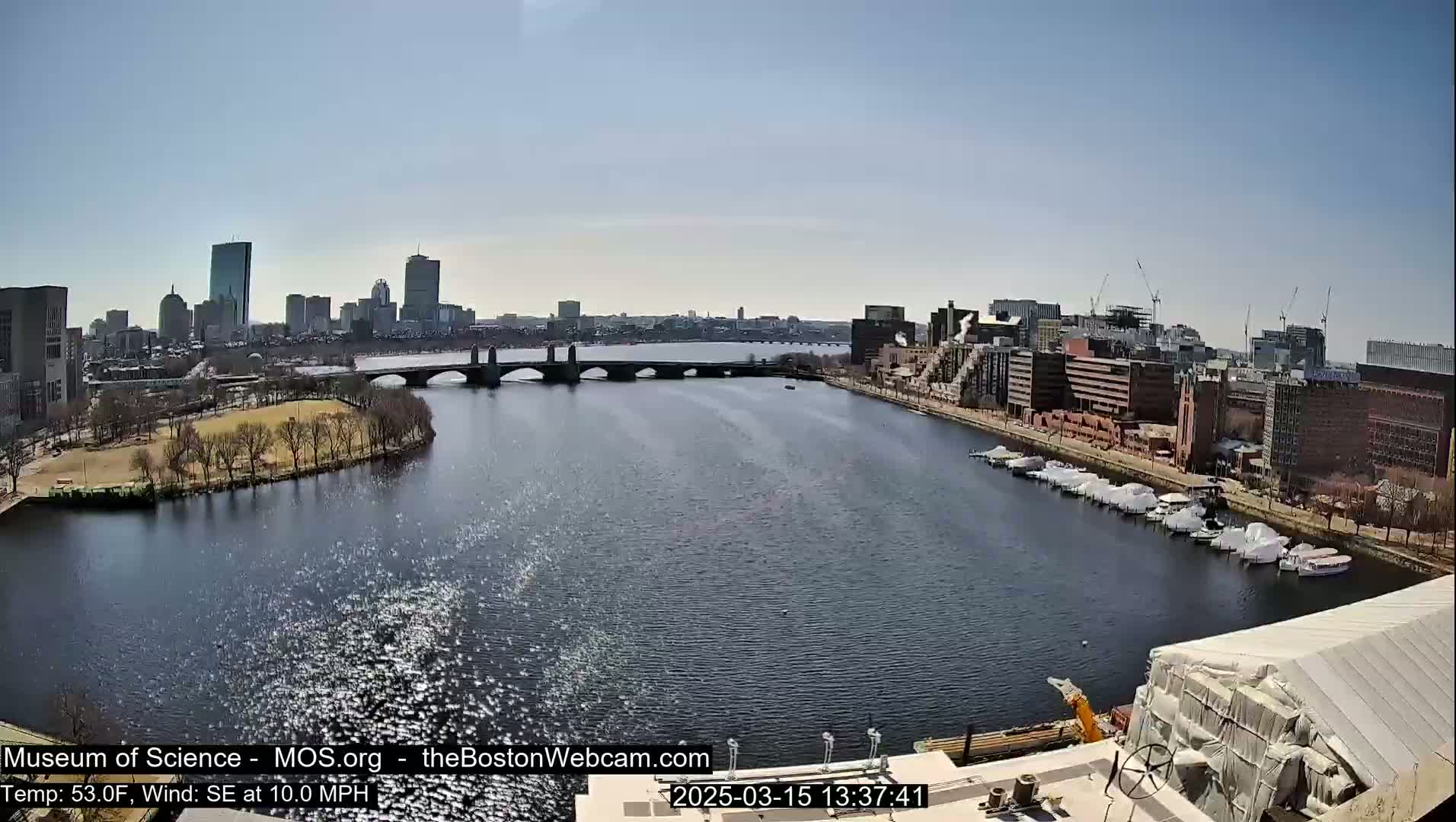 A sunny cityscape is viewed from above, showing a river with boats docked along its edge, a bridge spanning the river, and a cityscape in the background.