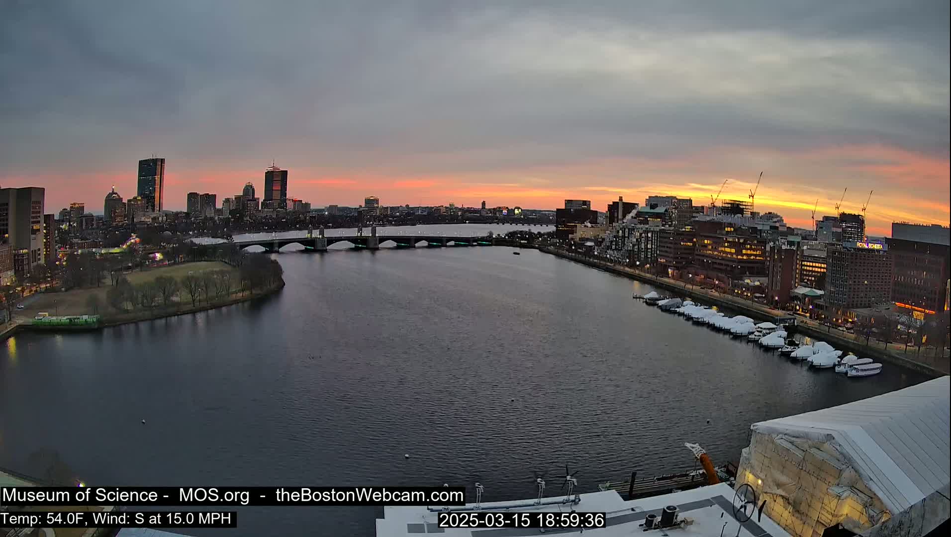 A city skyline at sunset over a river with a bridge, boats, and a park, under partly cloudy skies with a south wind of 15 mph.