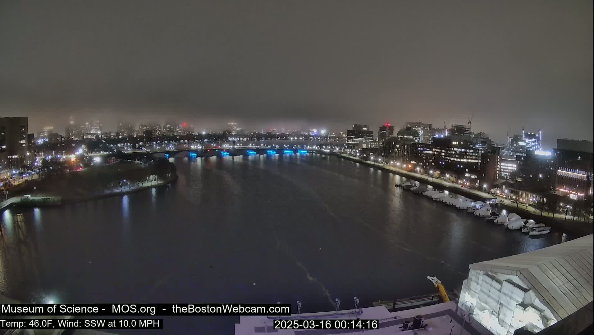 A nighttime, foggy view of a city waterfront with a river, bridges, buildings, and docked boats under a dark sky.