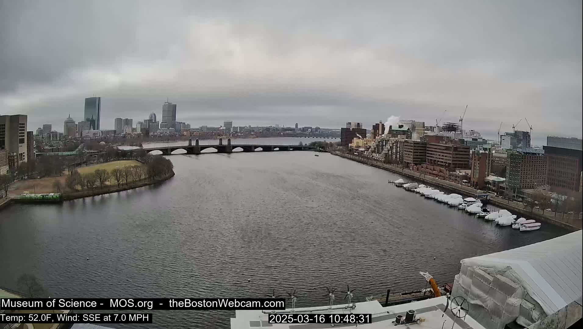 A city skyline with several buildings and a bridge over a river is visible under an overcast sky.
