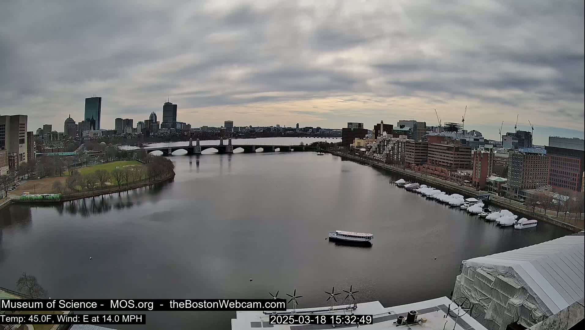 A city skyline is visible across a river under a cloudy sky, with a bridge, boats, and a partially covered building in the foreground.