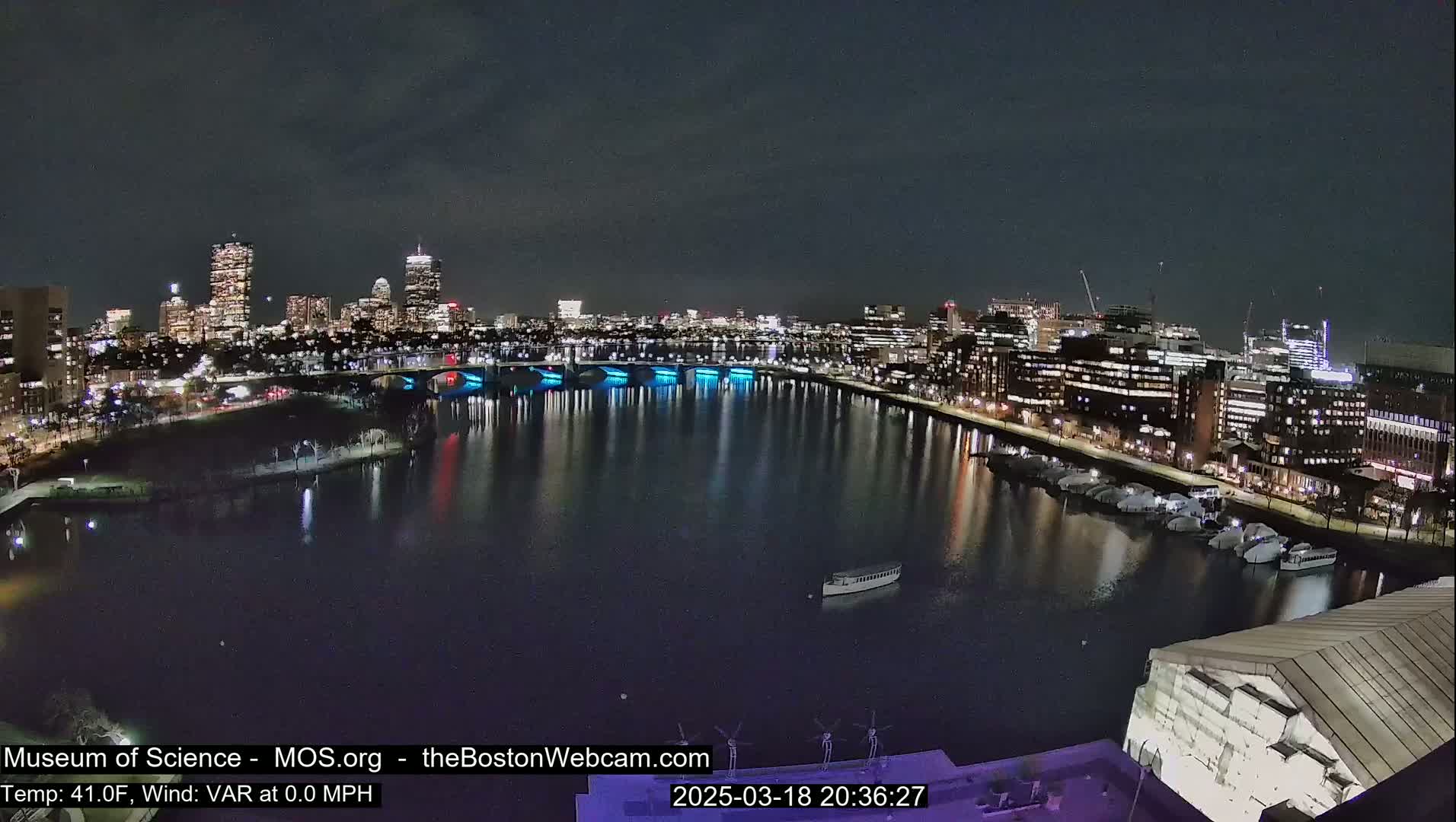 A nighttime cityscape with a calm body of water reflecting city lights, bridges, and boats under a clear, dark sky.