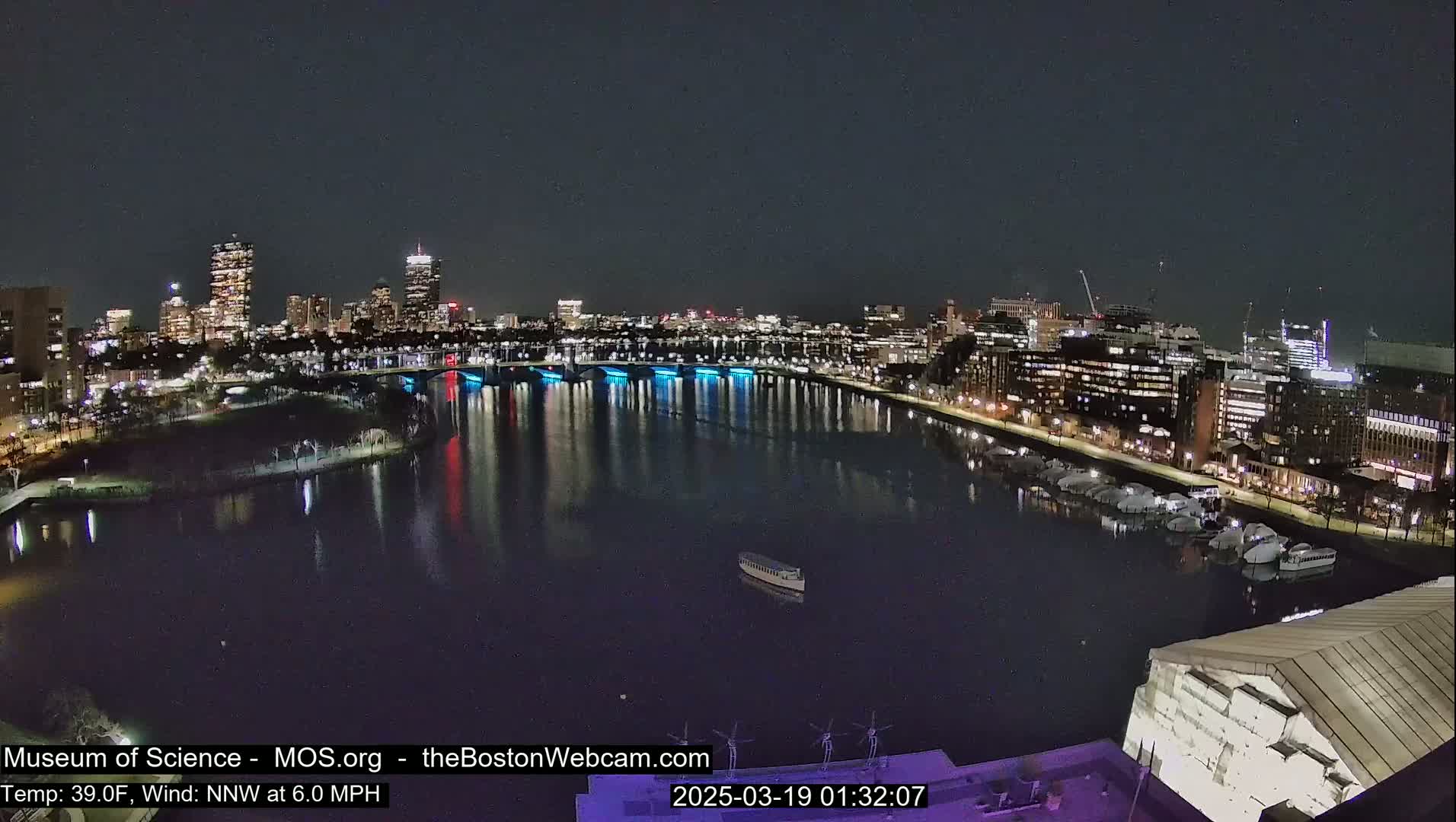 A nighttime view of a city skyline across a dark river, with a few boats docked and one in the middle of the river, under a clear sky.