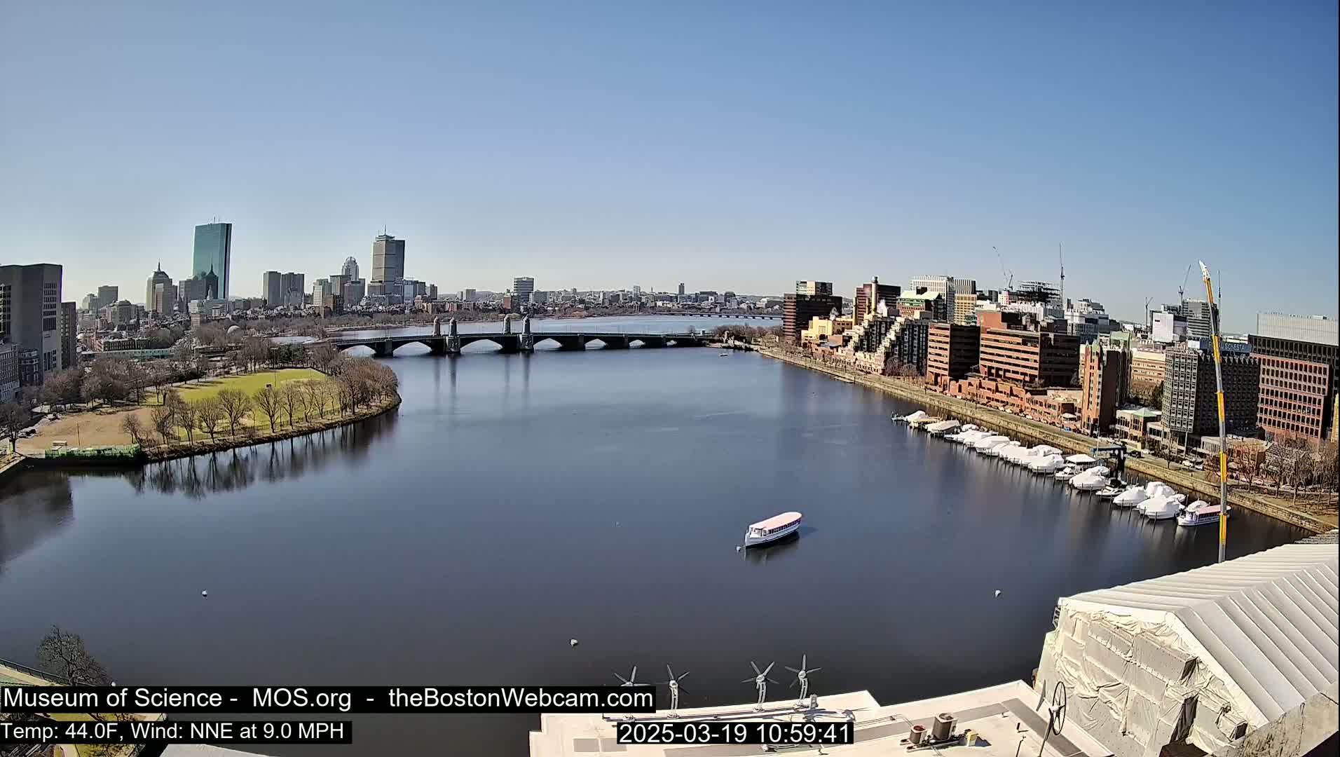 A calm river flows past a city skyline on a sunny day, with a bridge spanning the waterway and boats moored along the shore.