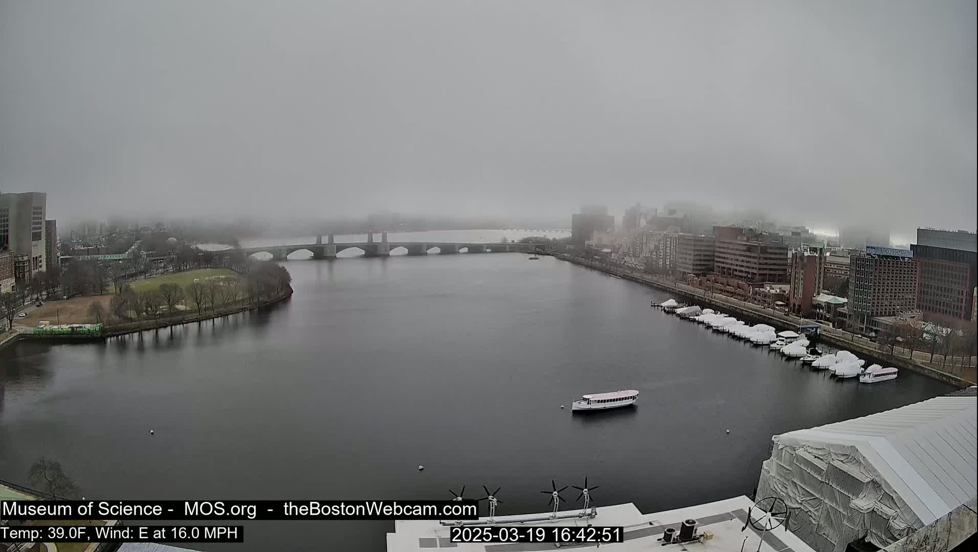 A foggy city scene shows a river with a bridge, boats, and buildings under a gray sky.
