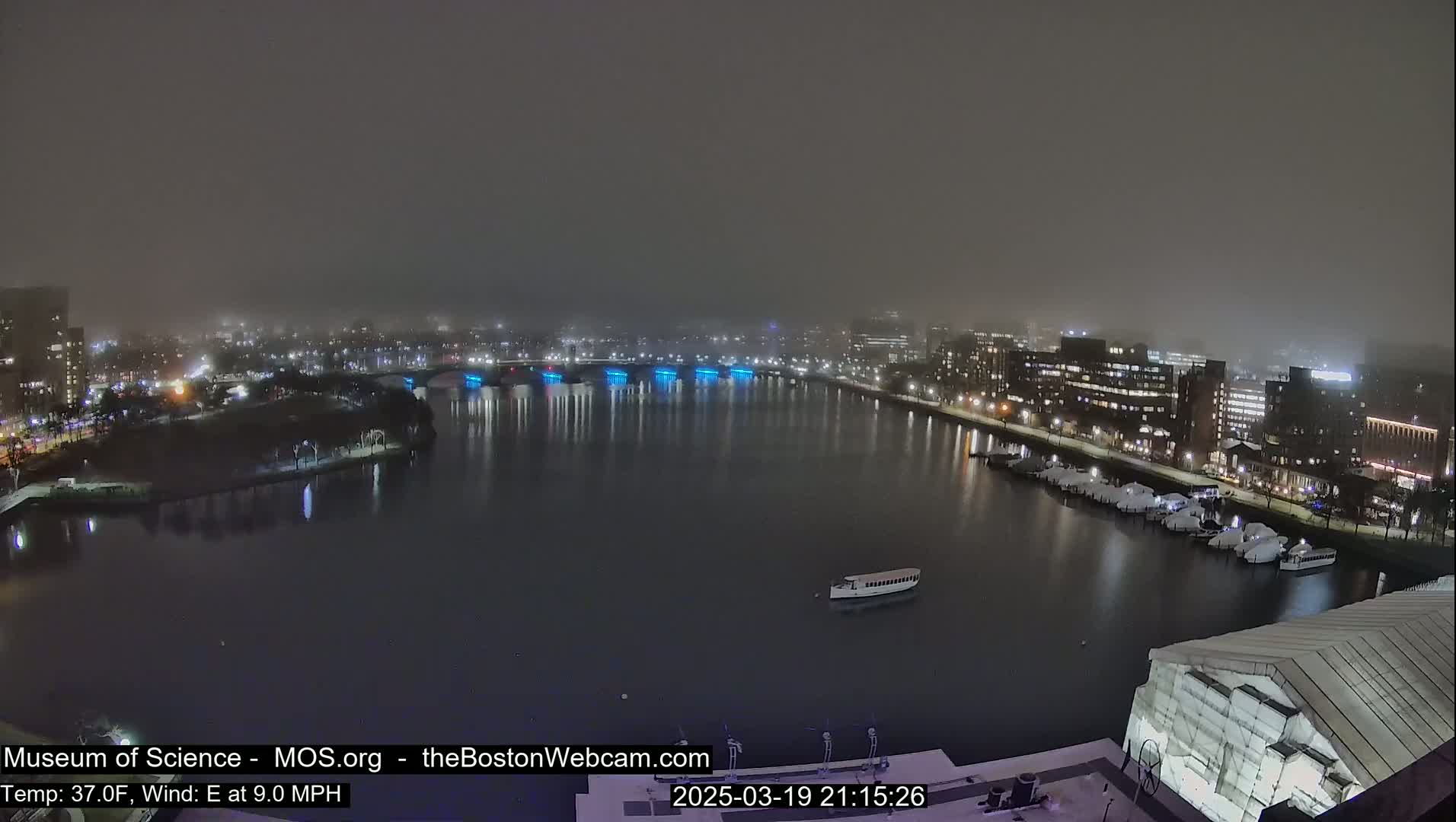 A nighttime, foggy view of a city waterfront with a bridge and docked boats, under a hazy sky.