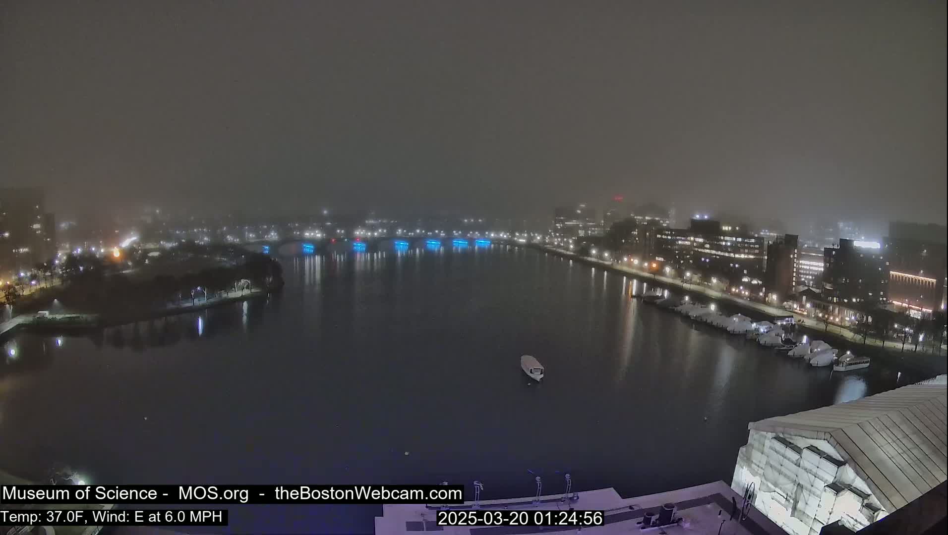A foggy night scene shows a body of water with city lights and docked boats, under a dark, hazy sky.