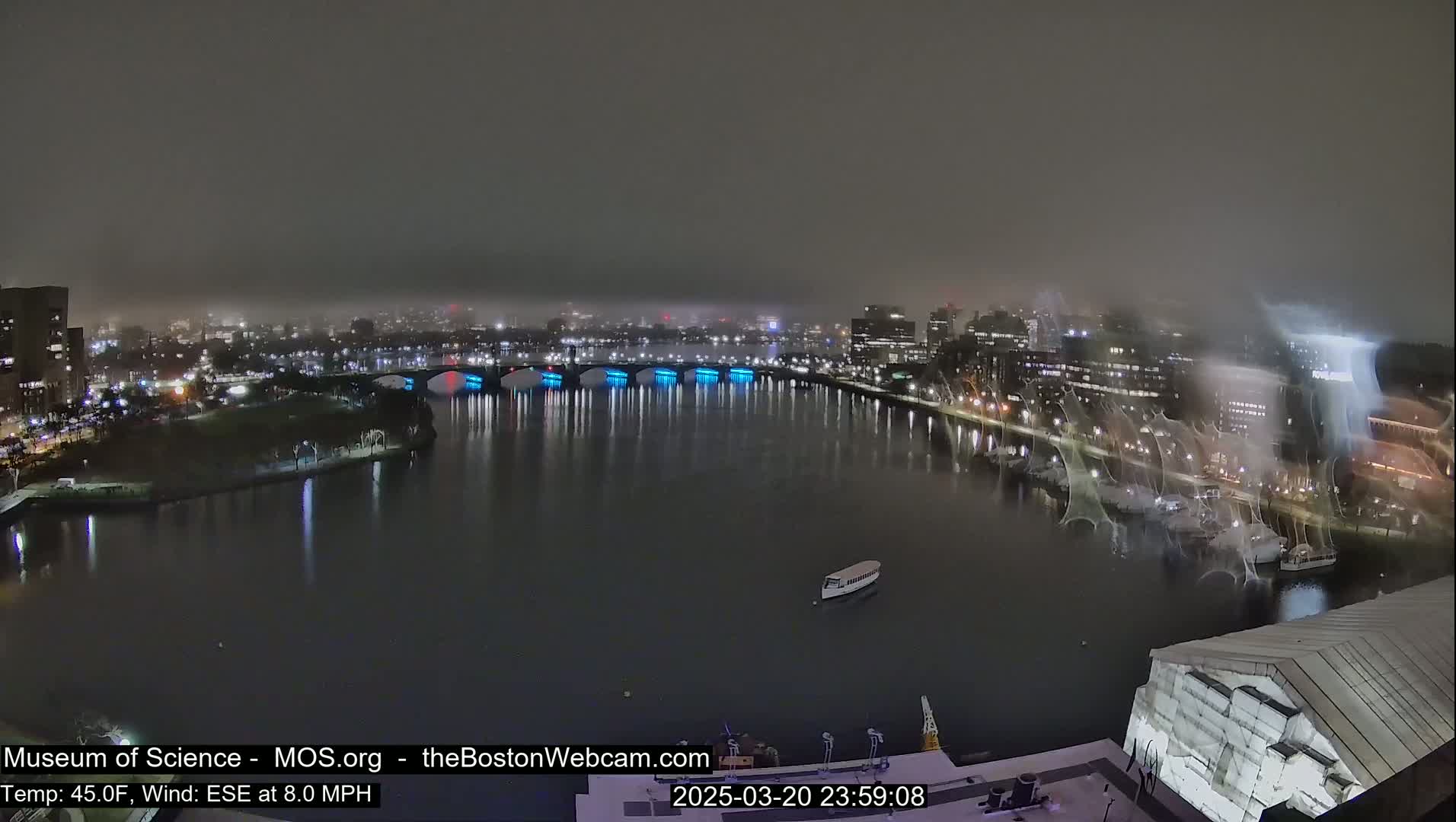 A nighttime view of a city skyline across a body of water, under foggy conditions, shows a bridge with blue lights and a few boats on the water.