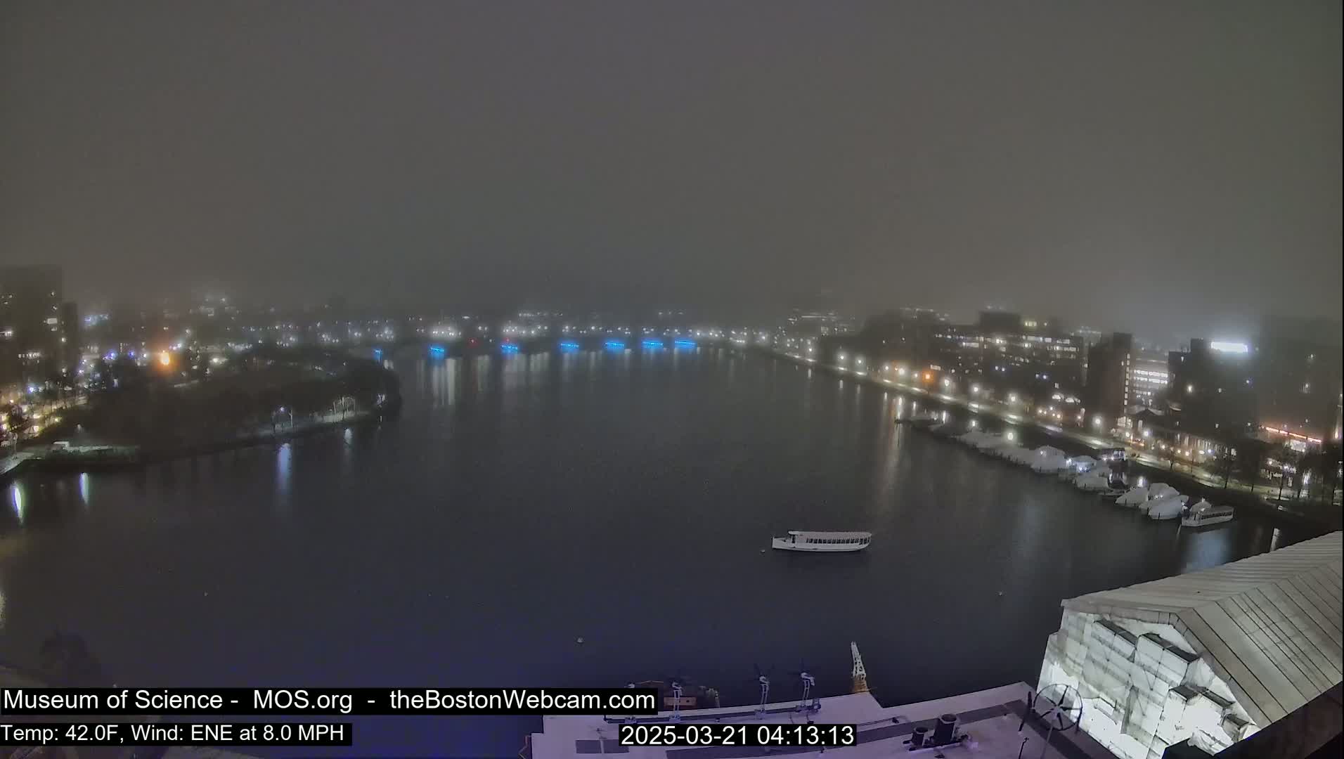 A foggy night scene shows a body of water with boats and city lights across the shore.