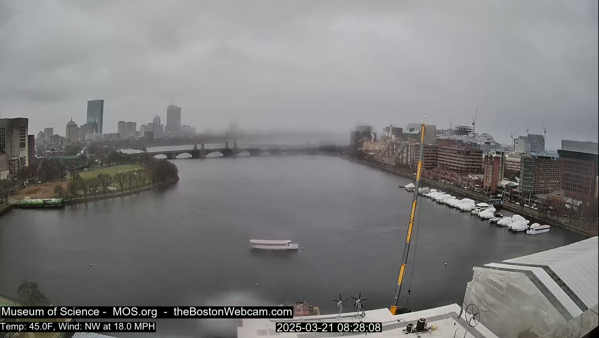 A foggy, overcast cityscape is seen across a wide river, with a bridge, boats, and docked yachts visible.