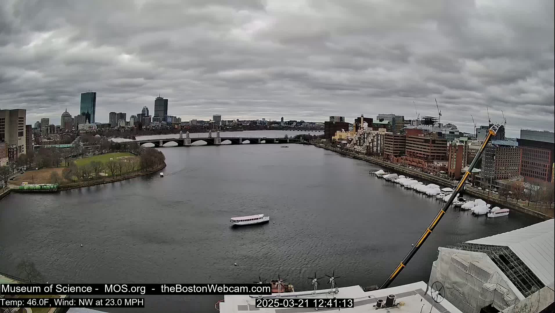 A wide shot of a city skyline across a river on an overcast day, showing a bridge, boats, and a construction crane.