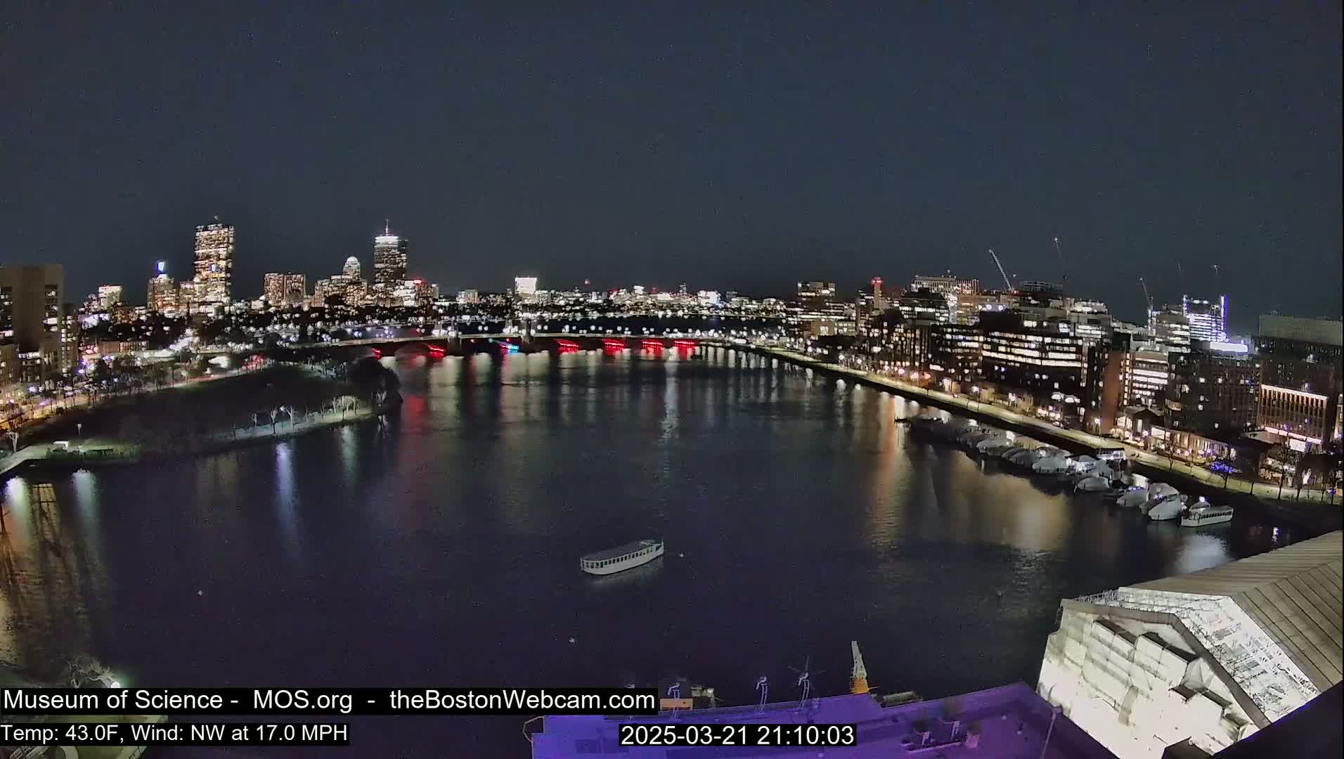 A nighttime view of a city skyline across a dark river, with boats moored along the waterfront under a clear sky.