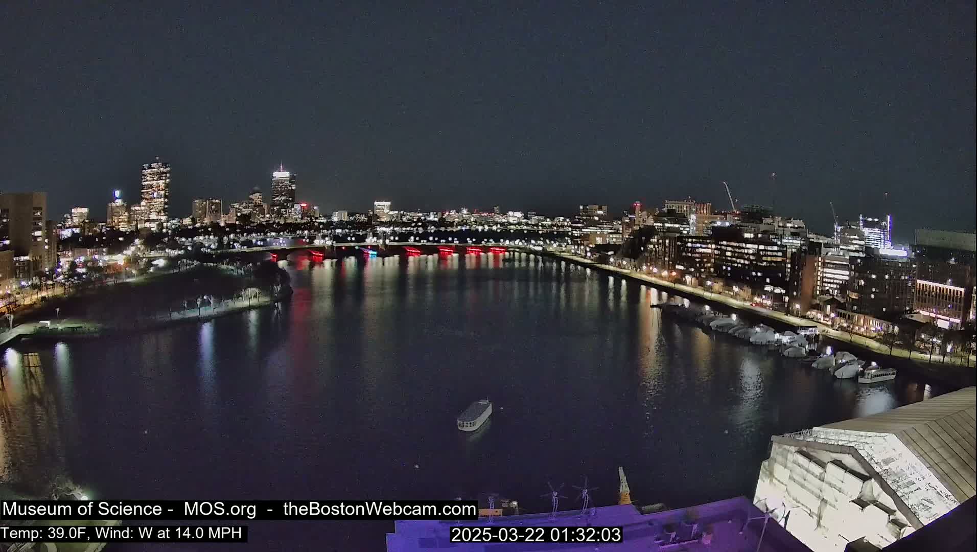 A nighttime view of a city skyline across a body of water with a west wind at 14 mph and a temperature of 39°F.