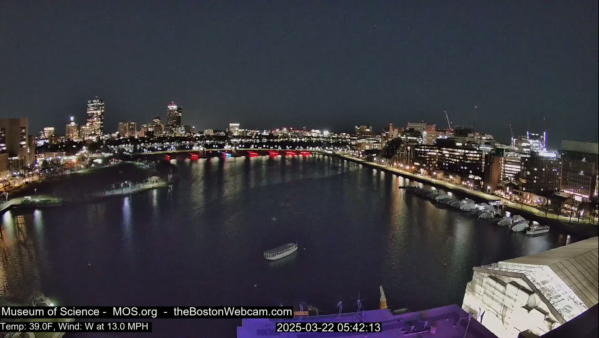 A nighttime view of a city skyline across a dark river with a moderate westerly wind and a temperature of 39°F.