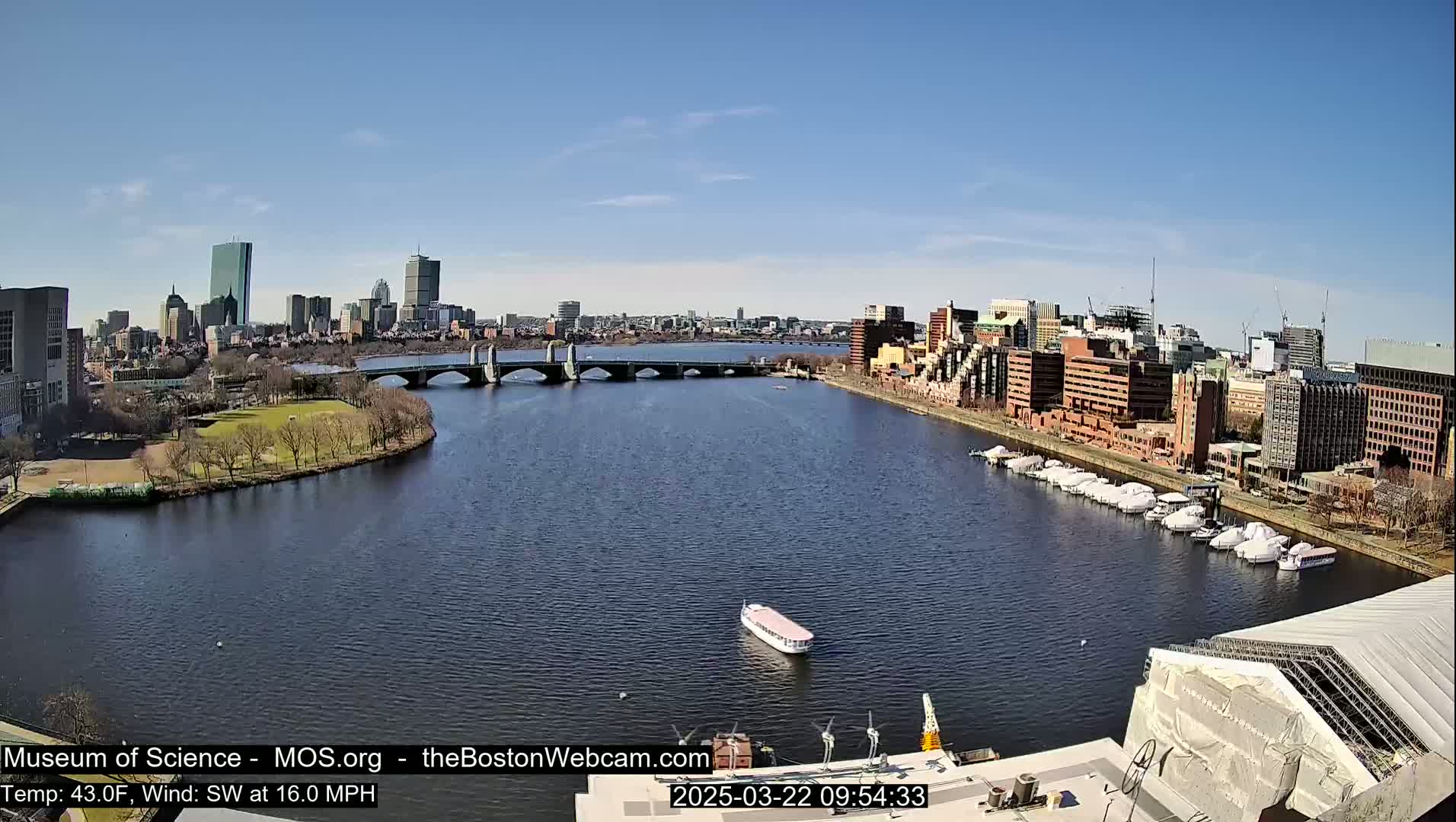 A river flows past a city skyline under a clear blue sky, with a bridge, boats, and a park visible on the shore.