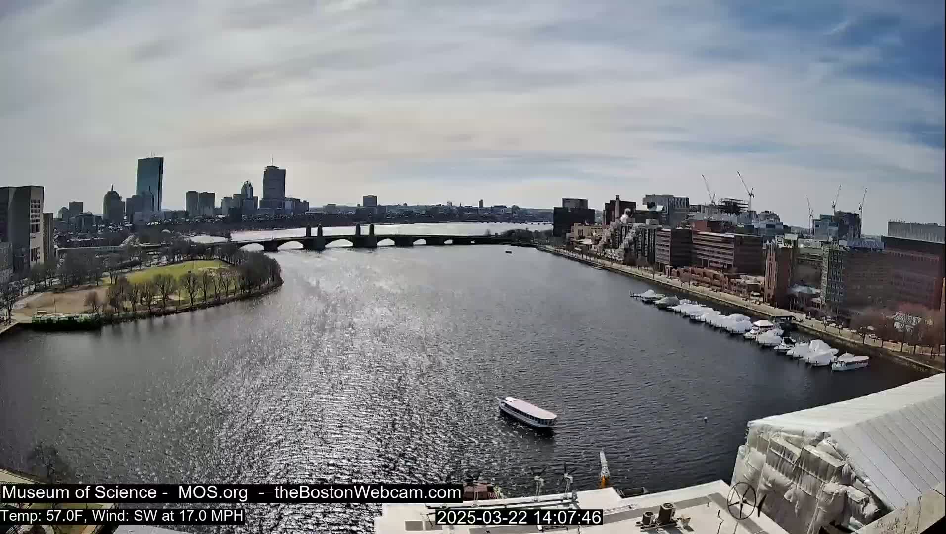A partly cloudy day shows a city skyline across a wide river with a bridge, boats, and docked vessels, a park, and some construction visible.