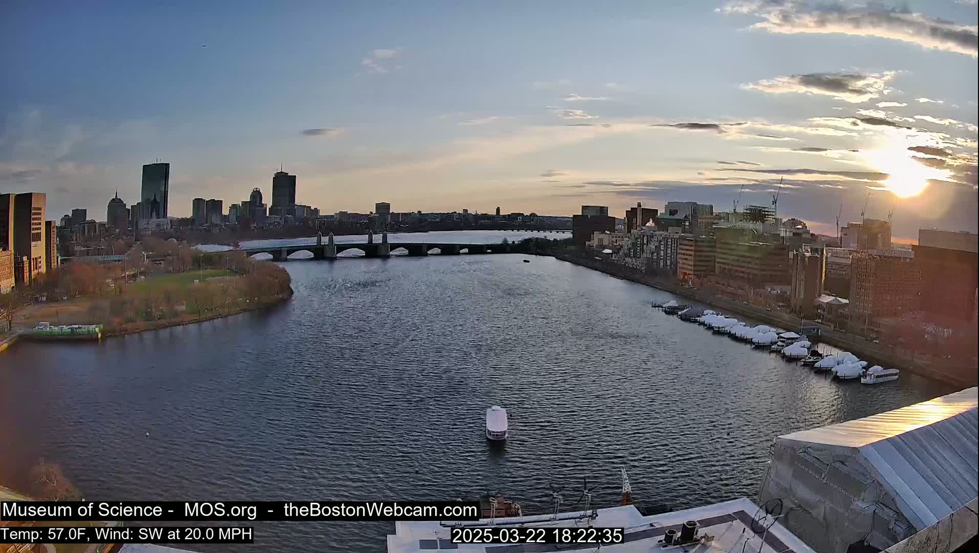 A wide shot of a city skyline at sunset over a river with a bridge, boats, and a calm, somewhat rippled body of water under a partly cloudy sky.