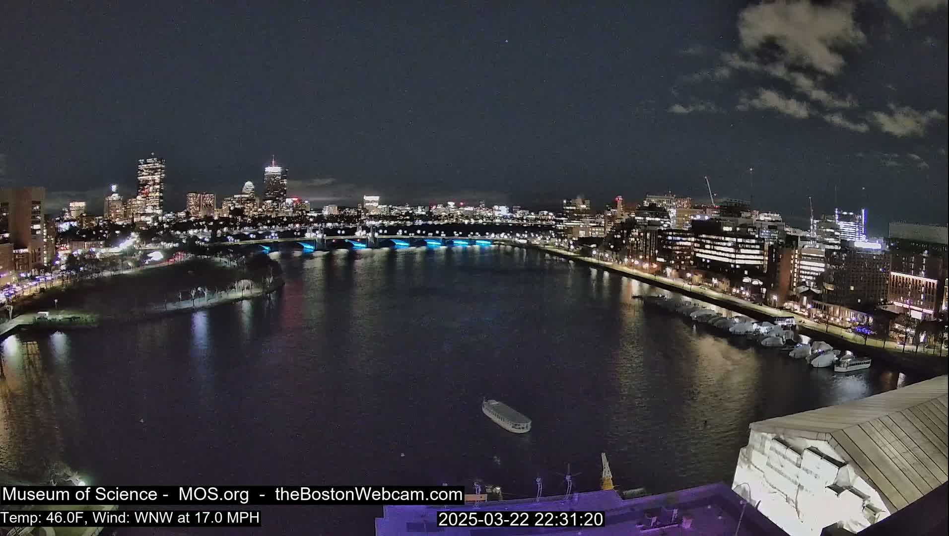 A nighttime view of a city skyline across a body of water, under a partly cloudy sky, with a boat on the water and numerous lights illuminating the city and waterfront.
