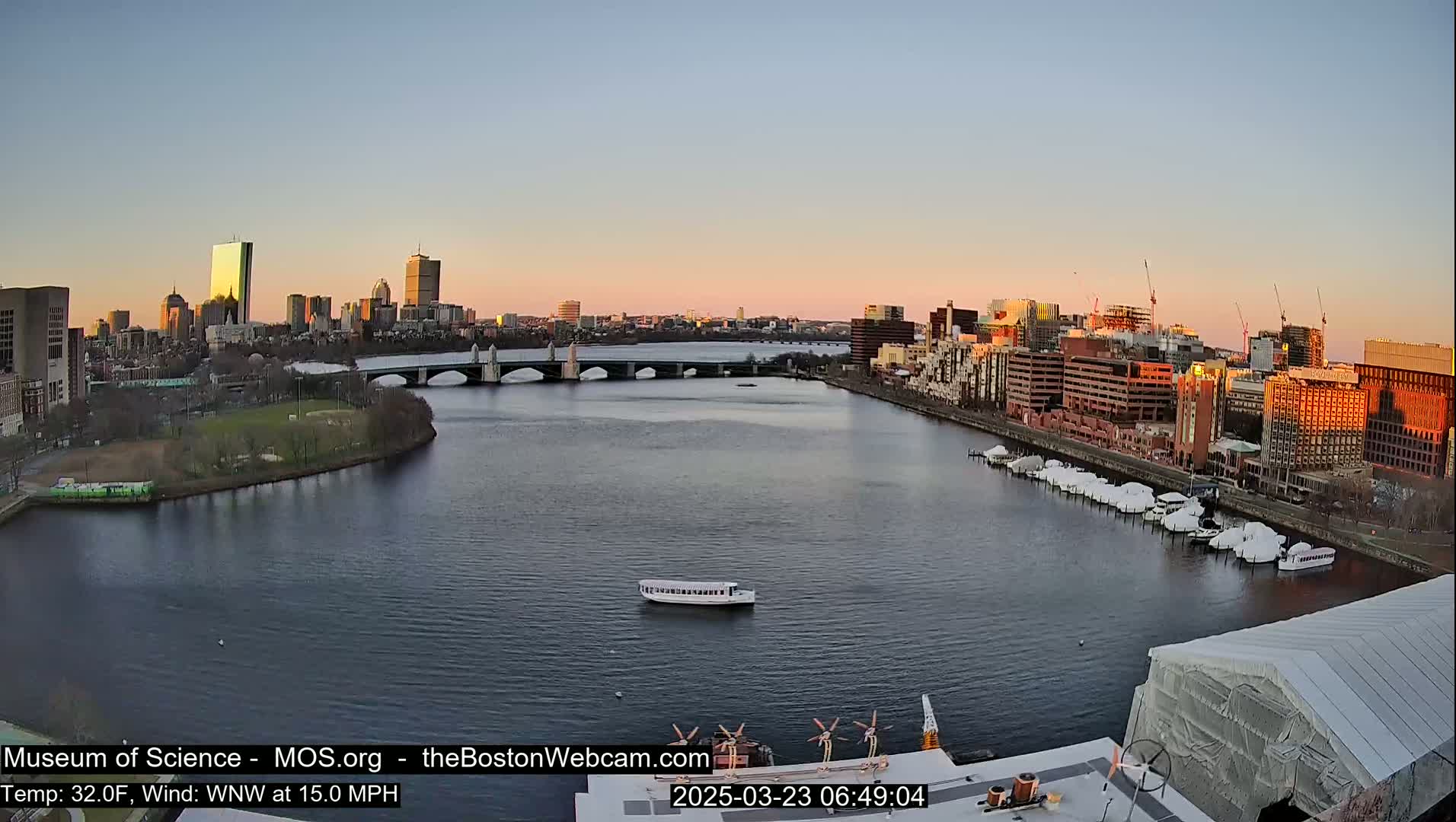 A city skyline at sunrise is viewed from across a calm river, with a bridge in the midground and numerous boats docked along the riverbank.