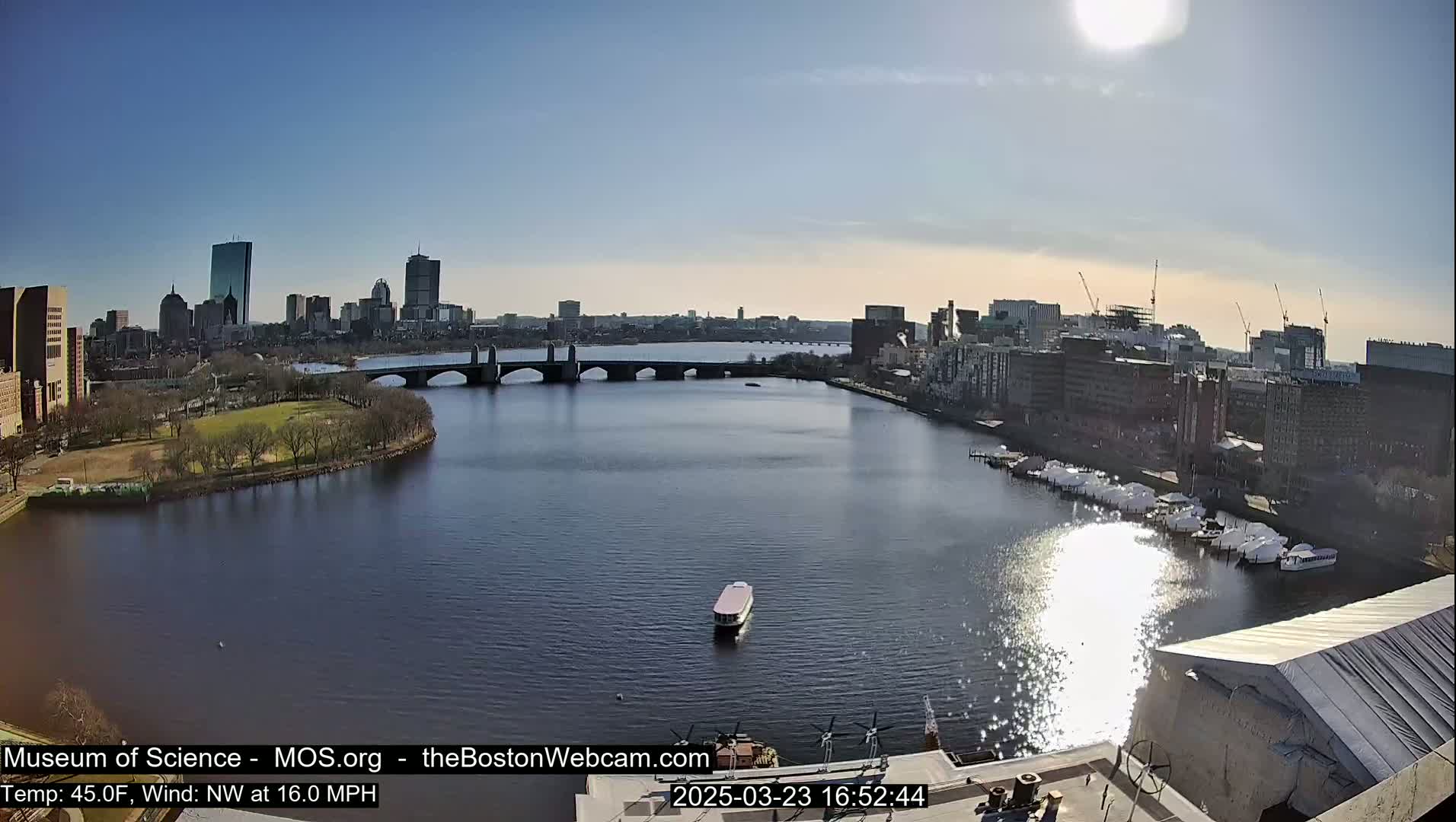 A sunny cityscape with a bridge over a calm river, boats docked along the shore, and a small vessel on the water.