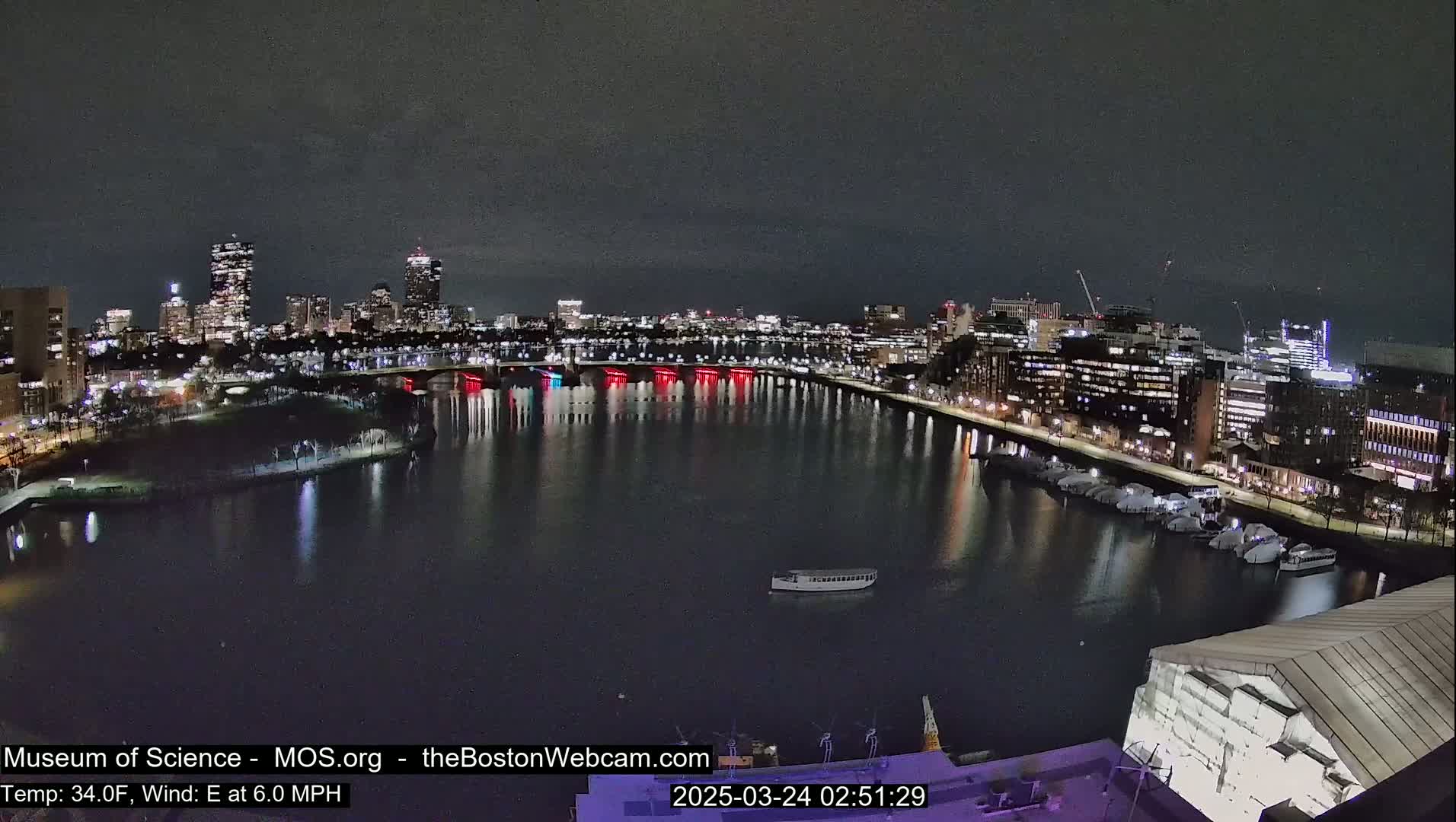 A nighttime view of a city skyline across a calm river, with a boat on the water and numerous lights illuminating buildings and bridges.