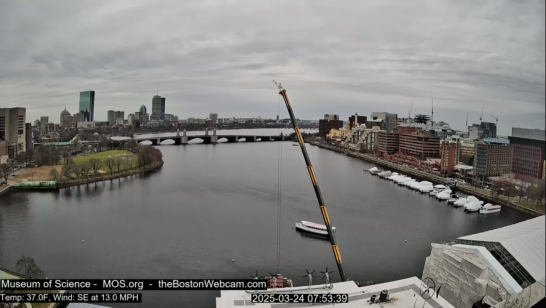 A large construction crane overlooks a river with a bridge and a city skyline under an overcast sky.