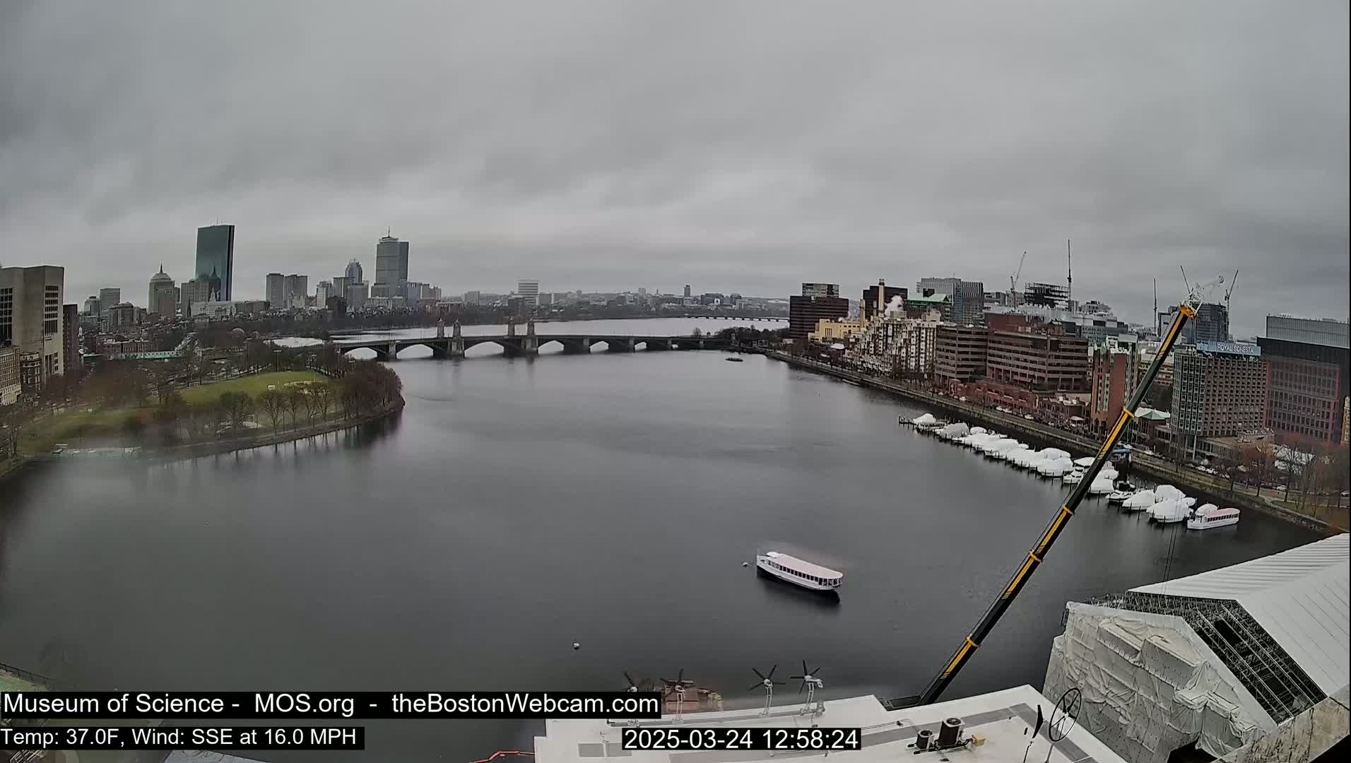 A city skyline with a bridge over a river, under an overcast sky; a boat is on the river, and a large crane is visible near a building under construction.