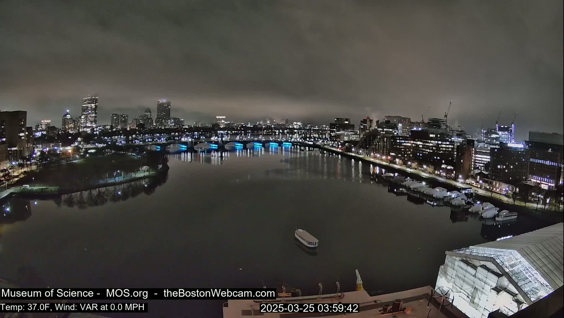 A nighttime view of a city skyline across a calm river, under a cloudy sky.