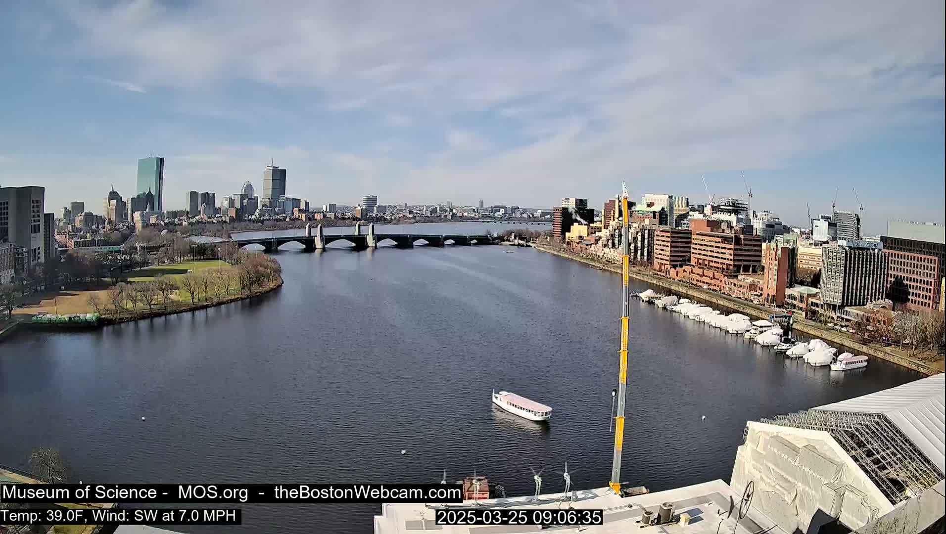 A river flows through a city under a partly cloudy sky, with a bridge spanning the river and boats docked along its banks.