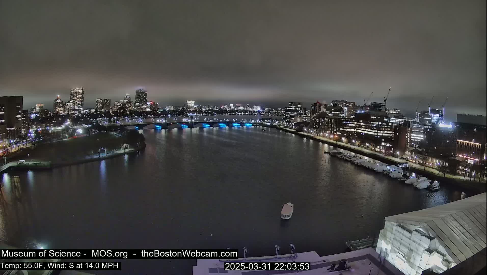 A nighttime cityscape is viewed across a dark river under an overcast sky, with a bridge illuminated in blue.