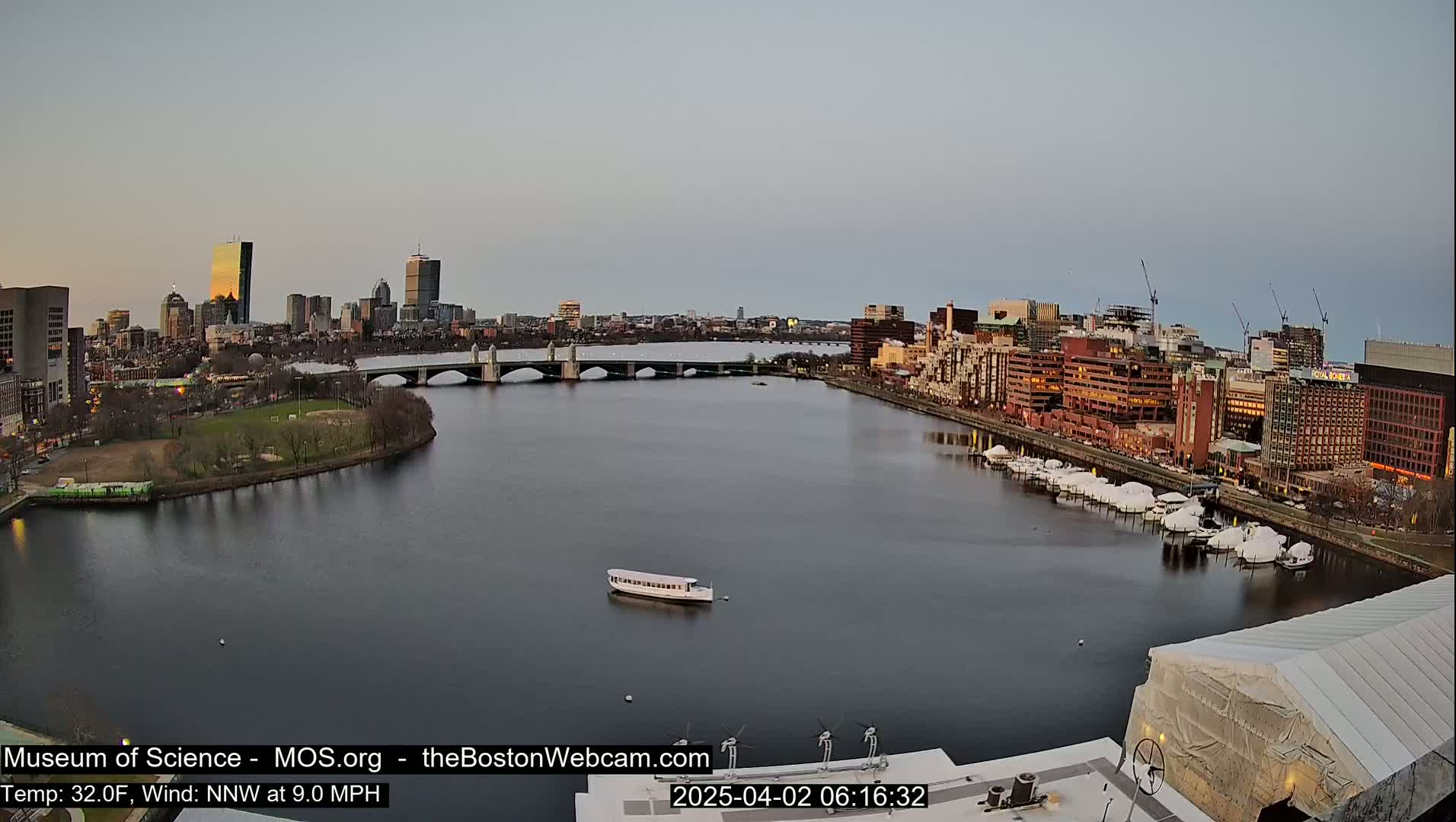 A calm river flows past a city skyline at dawn, with a bridge spanning the waterway and boats moored along the shore.