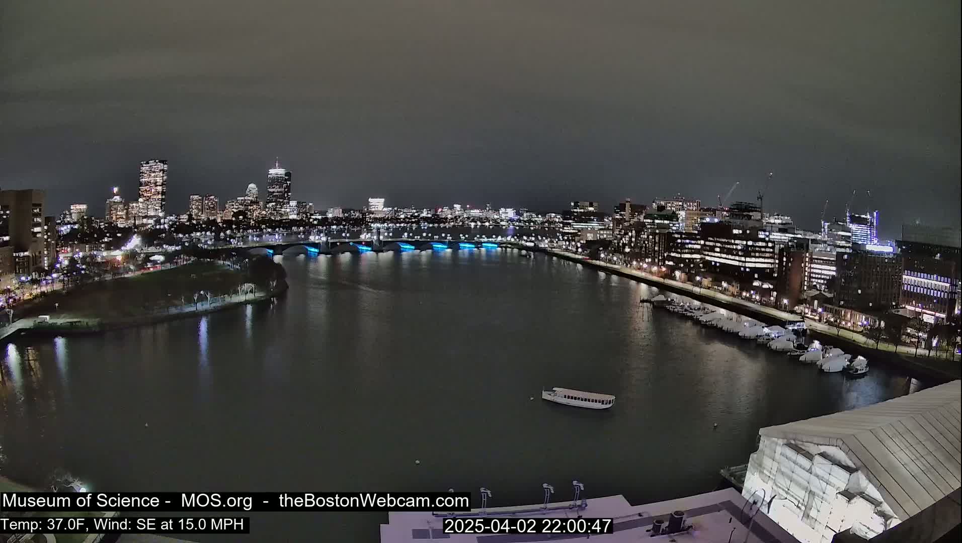 A nighttime cityscape across a calm river, with a bridge illuminated in blue, under a dark sky.