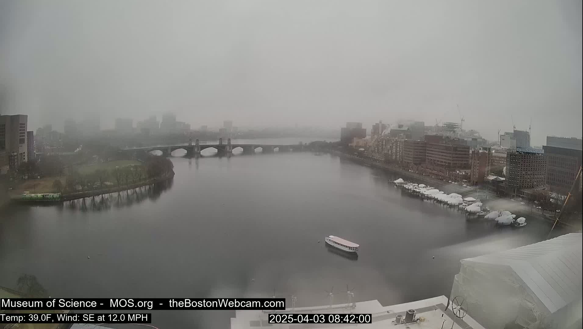 A foggy cityscape is seen from an elevated vantage point, showing a river with a bridge and docked boats, under a gray sky.