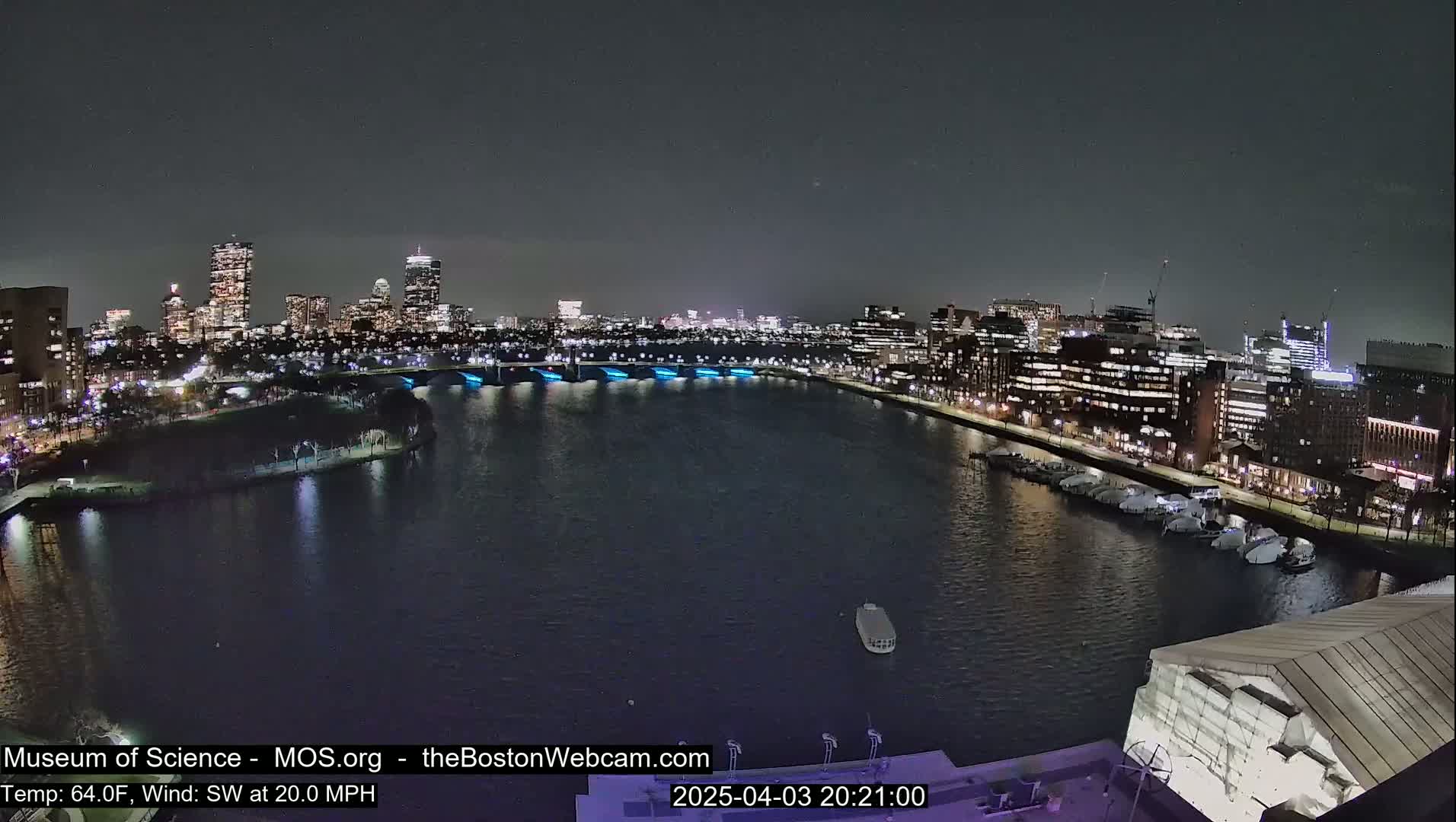 A nighttime cityscape across a body of water, with a city skyline illuminated, under a dark sky and a southwest wind of 20 mph.