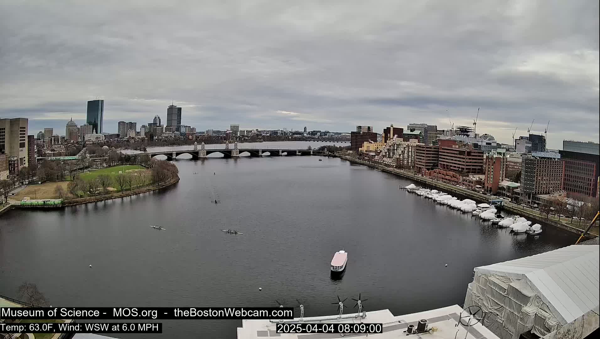 A wide shot of a river under an overcast sky shows a city skyline in the distance, a bridge, several boats, and a few rowers.