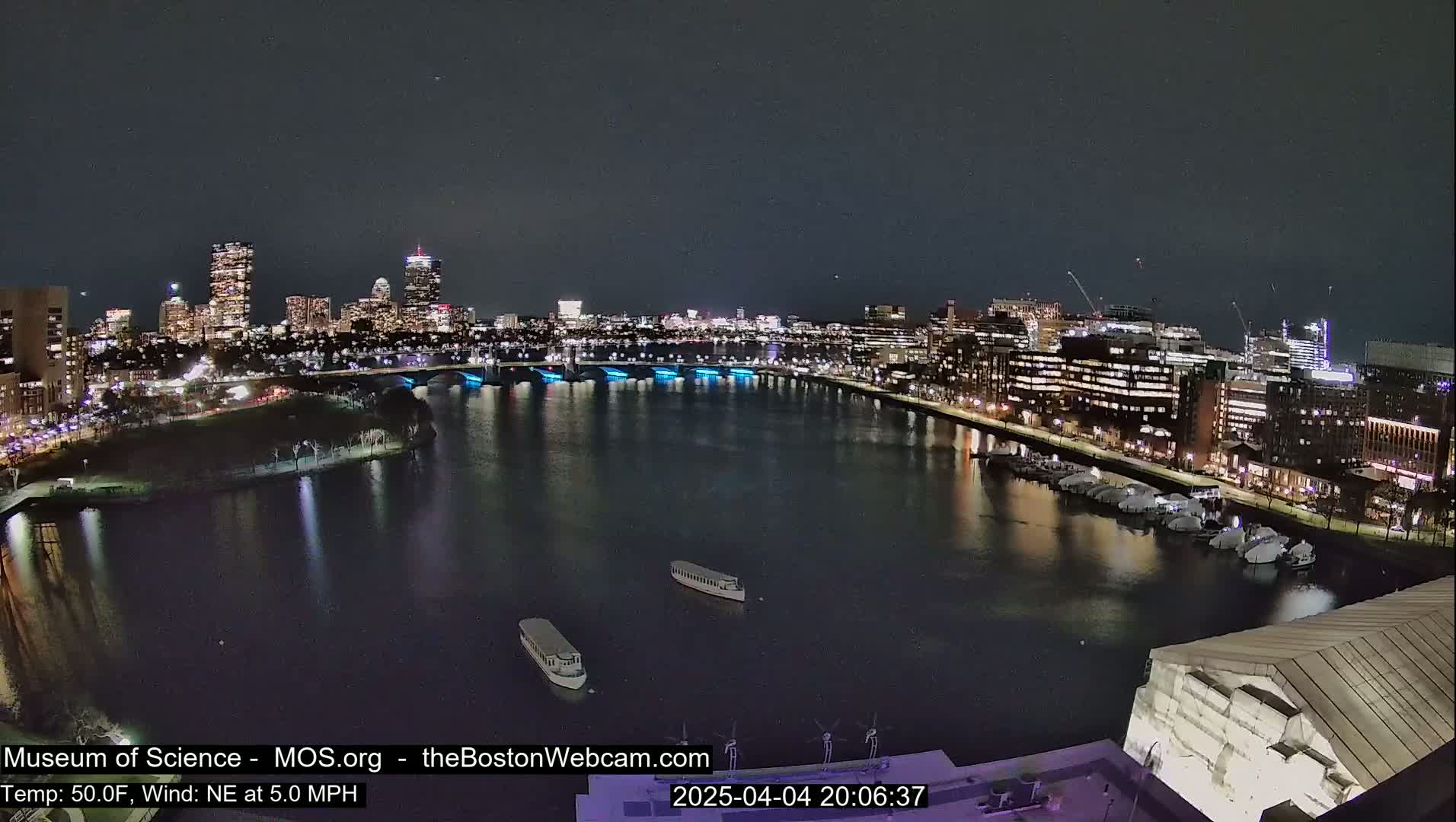 A nighttime view of a city skyline across a calm body of water, with boats docked along the shore and a bridge illuminated with blue lights in the background, under a clear night sky.