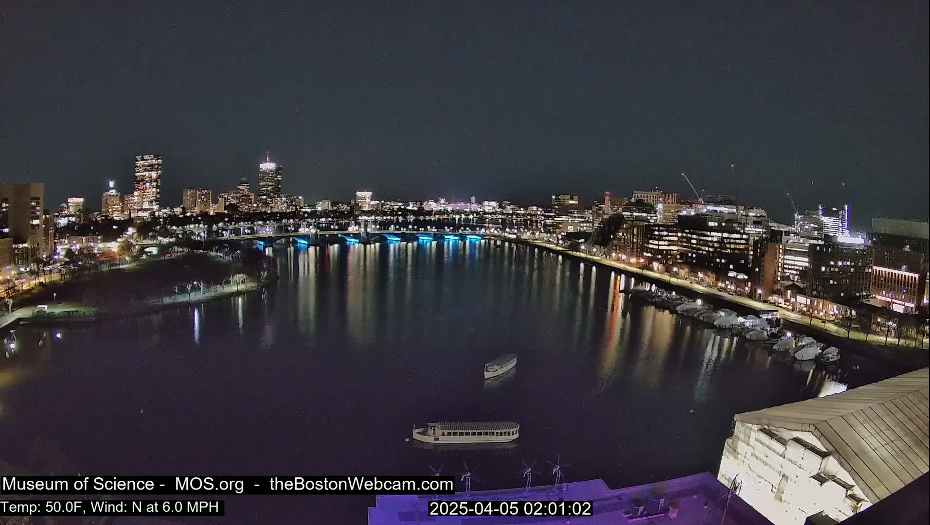 A nighttime view of a city skyline across a calm body of water, with boats docked along the shore under a clear, dark sky.