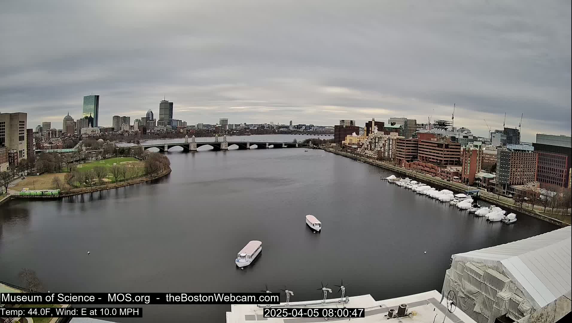 A wide shot shows a city skyline across a river with a bridge, boats docked along the shore, and two tour boats on the water under an overcast sky.