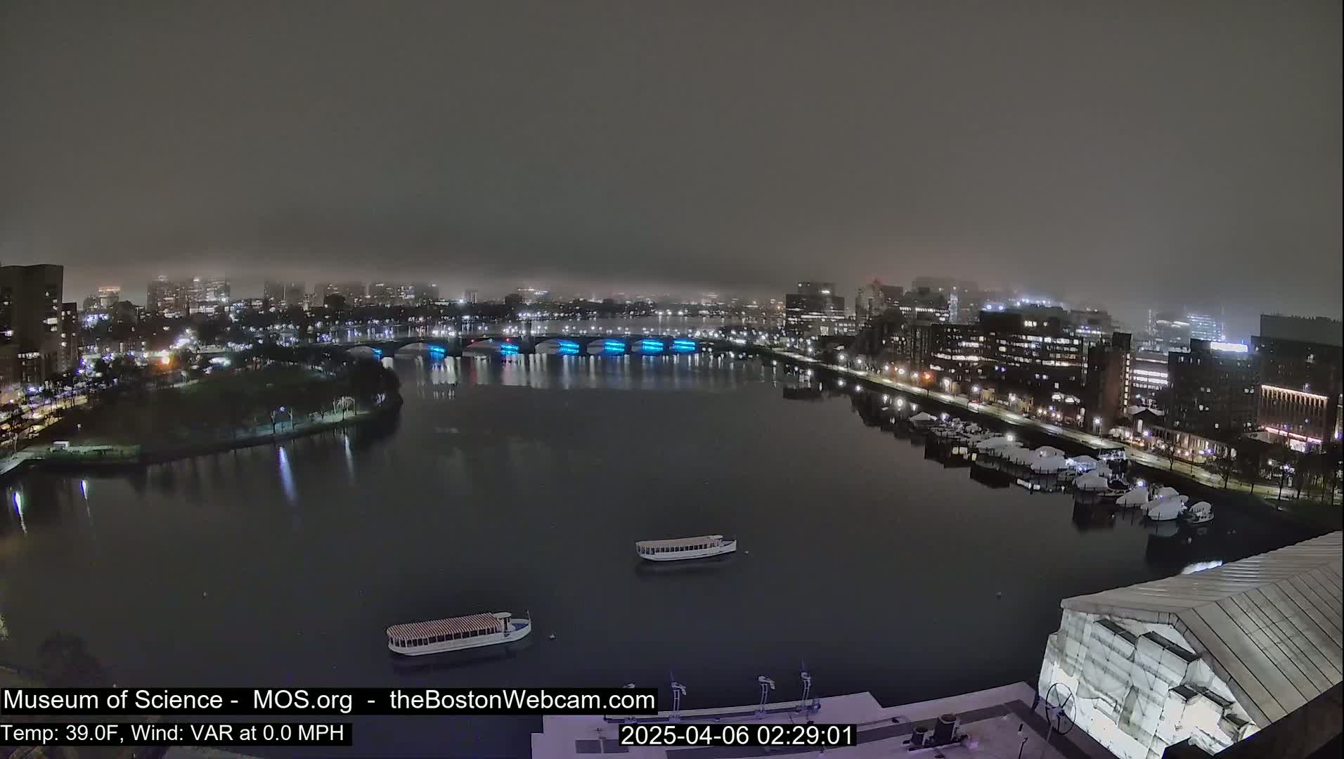 A nighttime view of a city waterfront shrouded in fog, showing a river with boats and lit buildings along the shore.