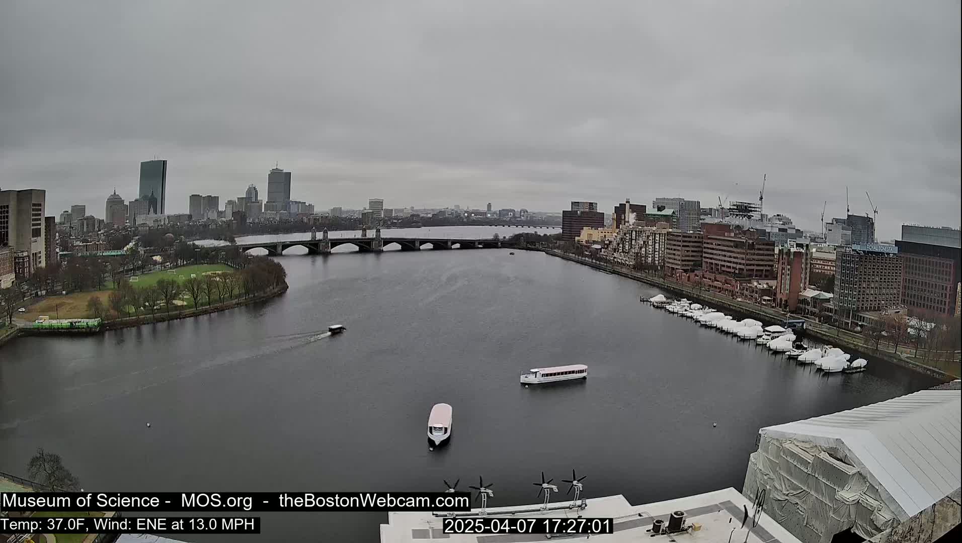 A wide shot of a city skyline across a river on an overcast day, with boats moored on the right bank and a few small boats on the water.