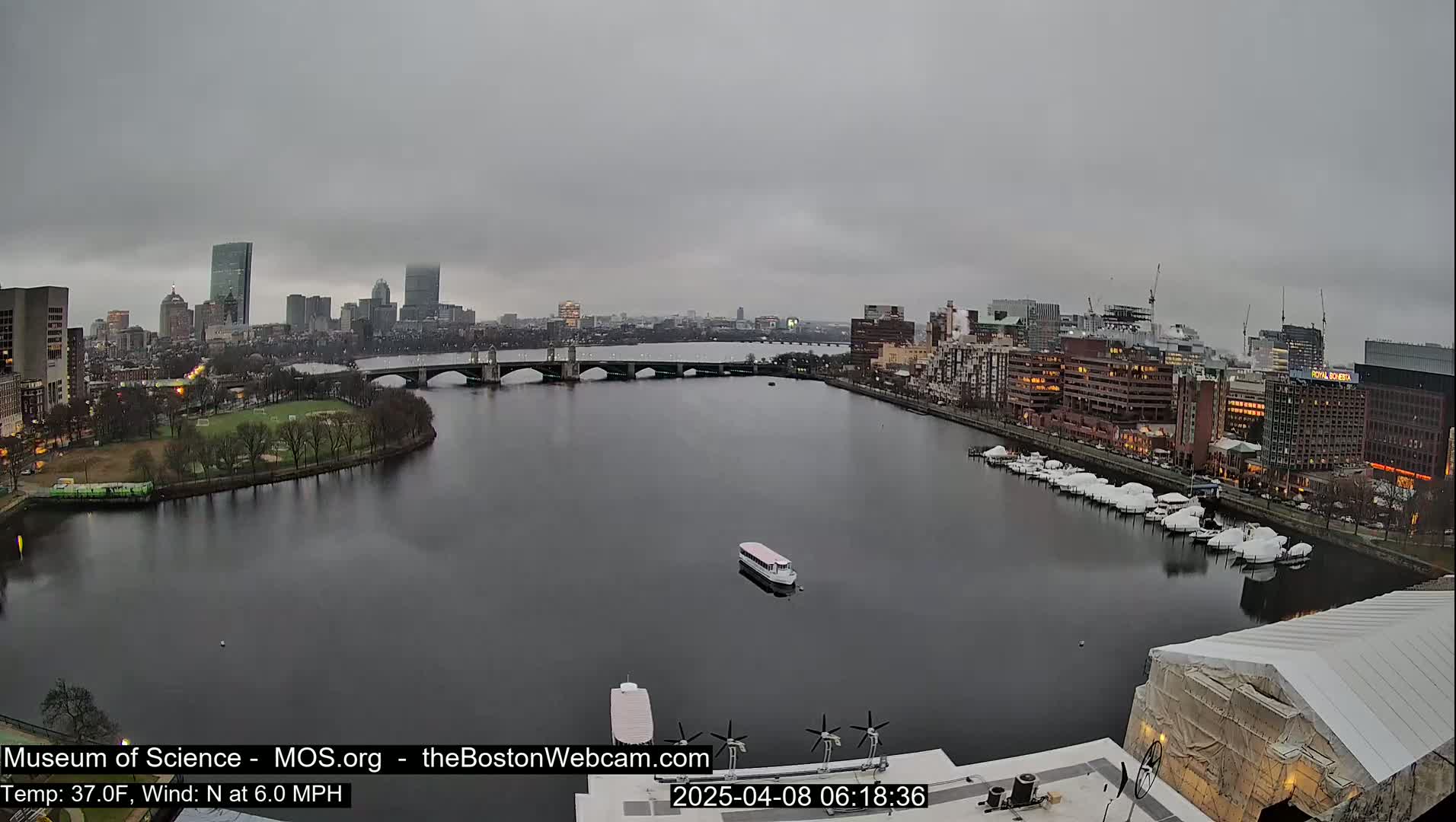 A city skyline under an overcast sky is visible across a calm river, with a bridge and docked boats in the foreground.