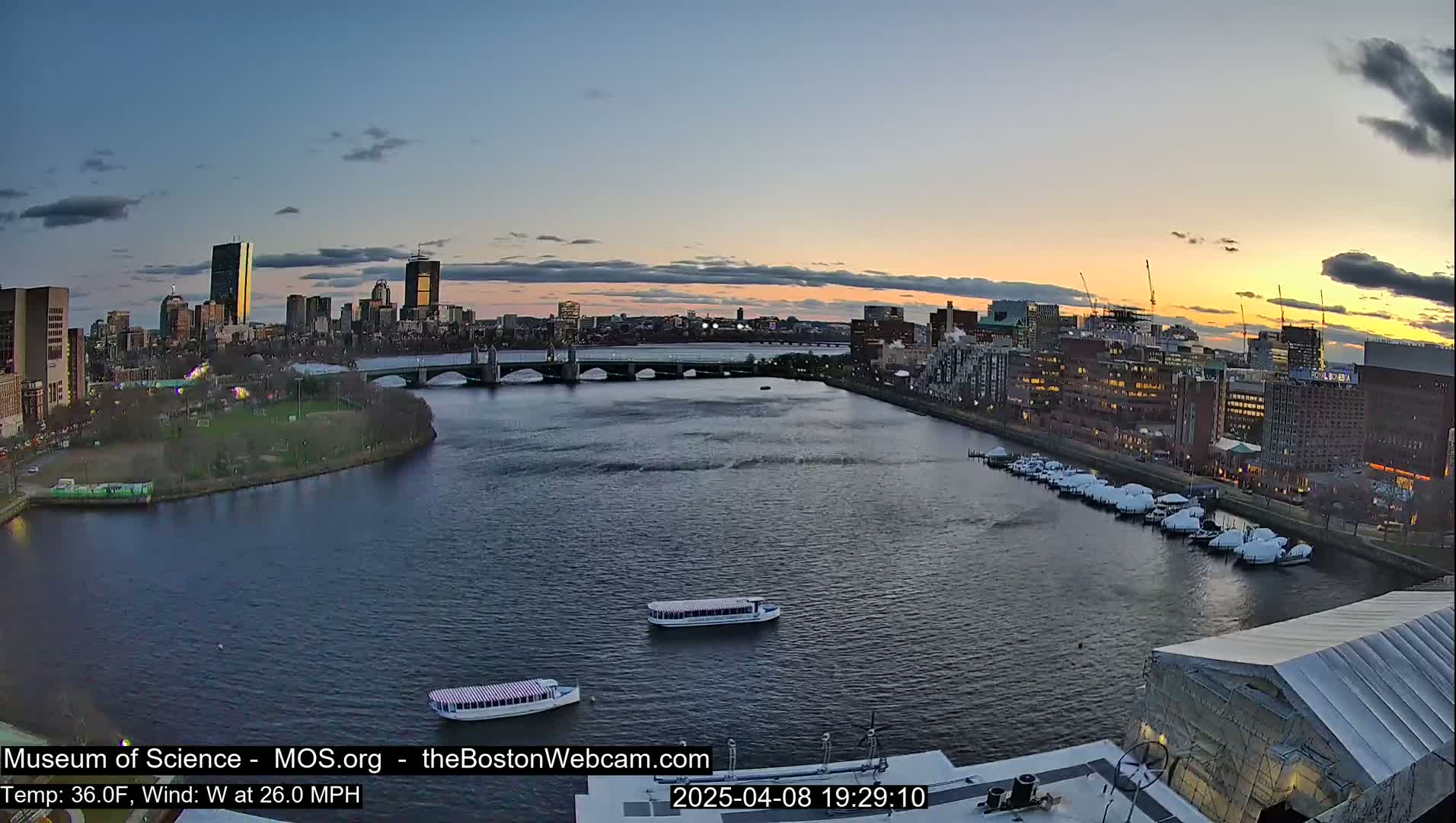 A city skyline at sunset is visible across a river with two tour boats and a marina, under a mostly clear sky with a light wind.