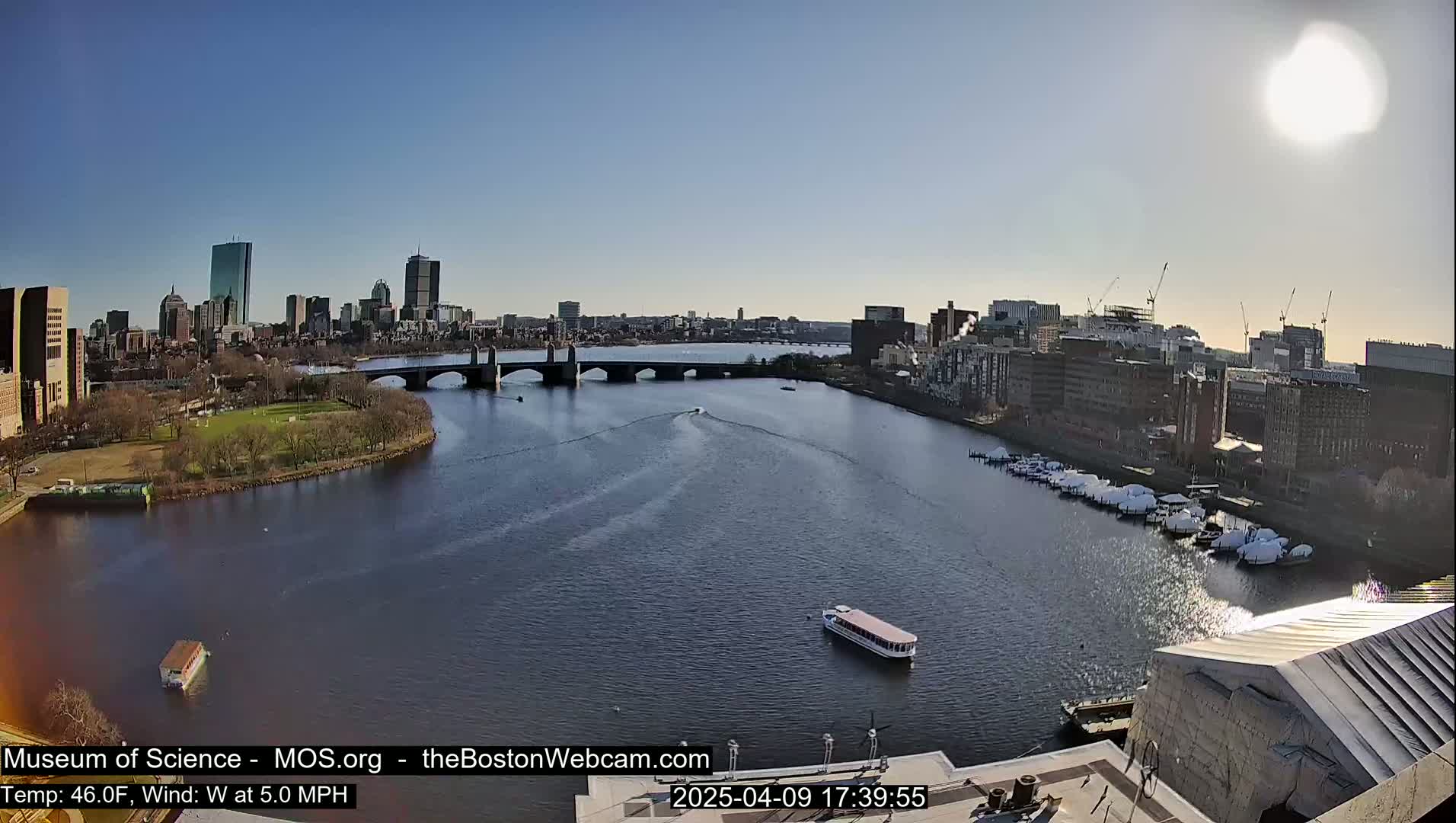 A sunny cityscape is viewed from above, showing a river with boats and a bridge, with a city skyline in the background.