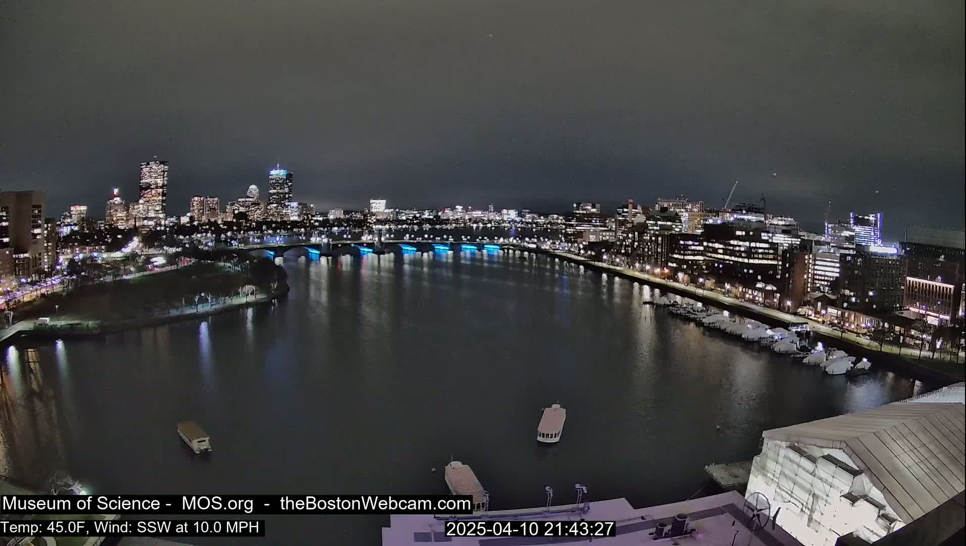 A nighttime view of a city skyline across a dark river, with boats on the water and a building under construction in the foreground, under a dark, clear sky with a 10 mph south-southwest wind and a temperature of 45°F.