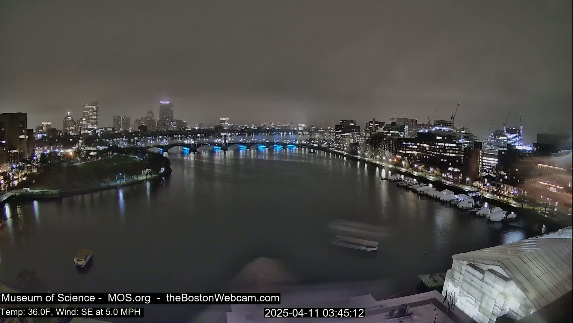 A nighttime view of a city skyline across a river under a foggy sky, with boats on the water and lights illuminating the bridges and buildings.