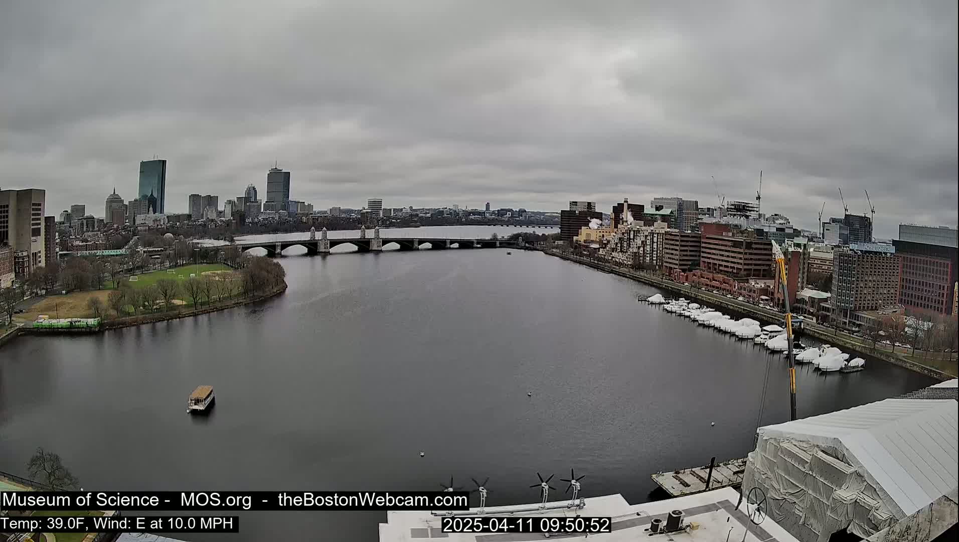 A river flows through a city under an overcast sky, with a bridge spanning the river and boats moored along its banks.