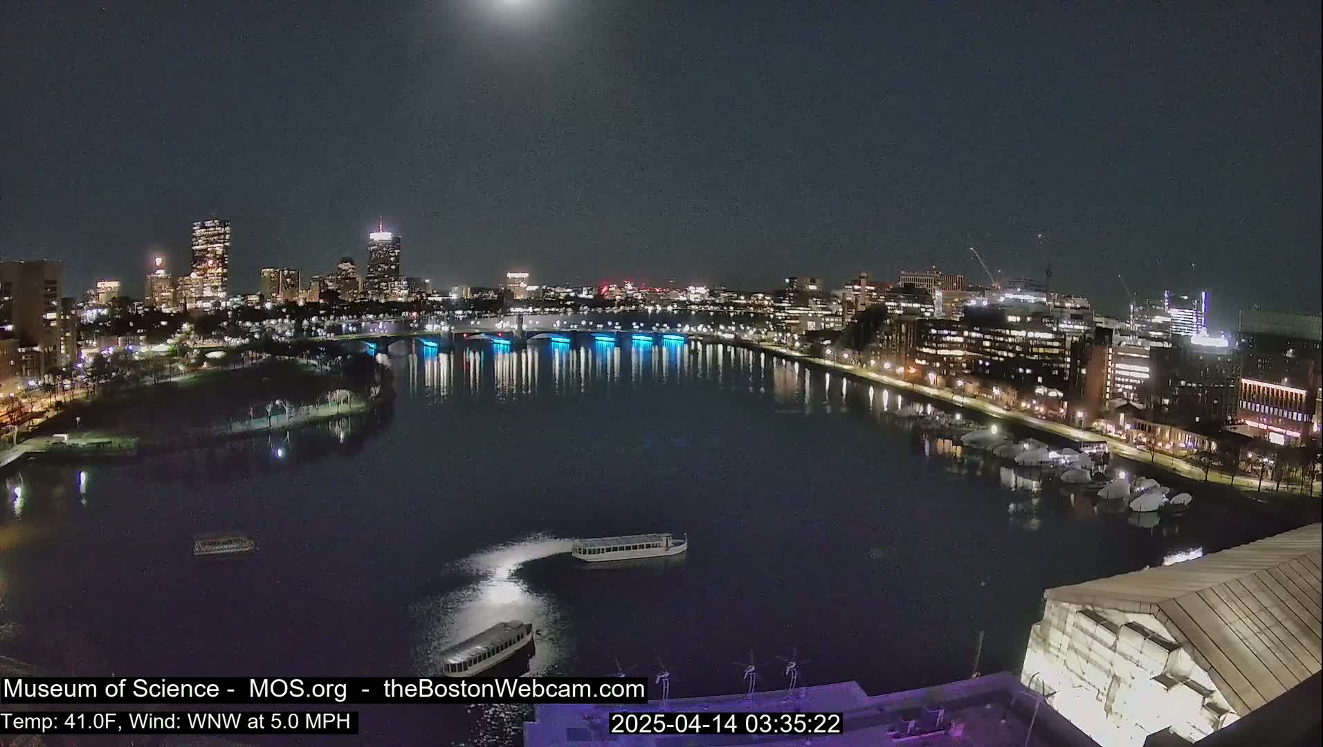 A nighttime cityscape with a moonlit river reflecting city lights, several boats on the water, and a calm, clear sky.