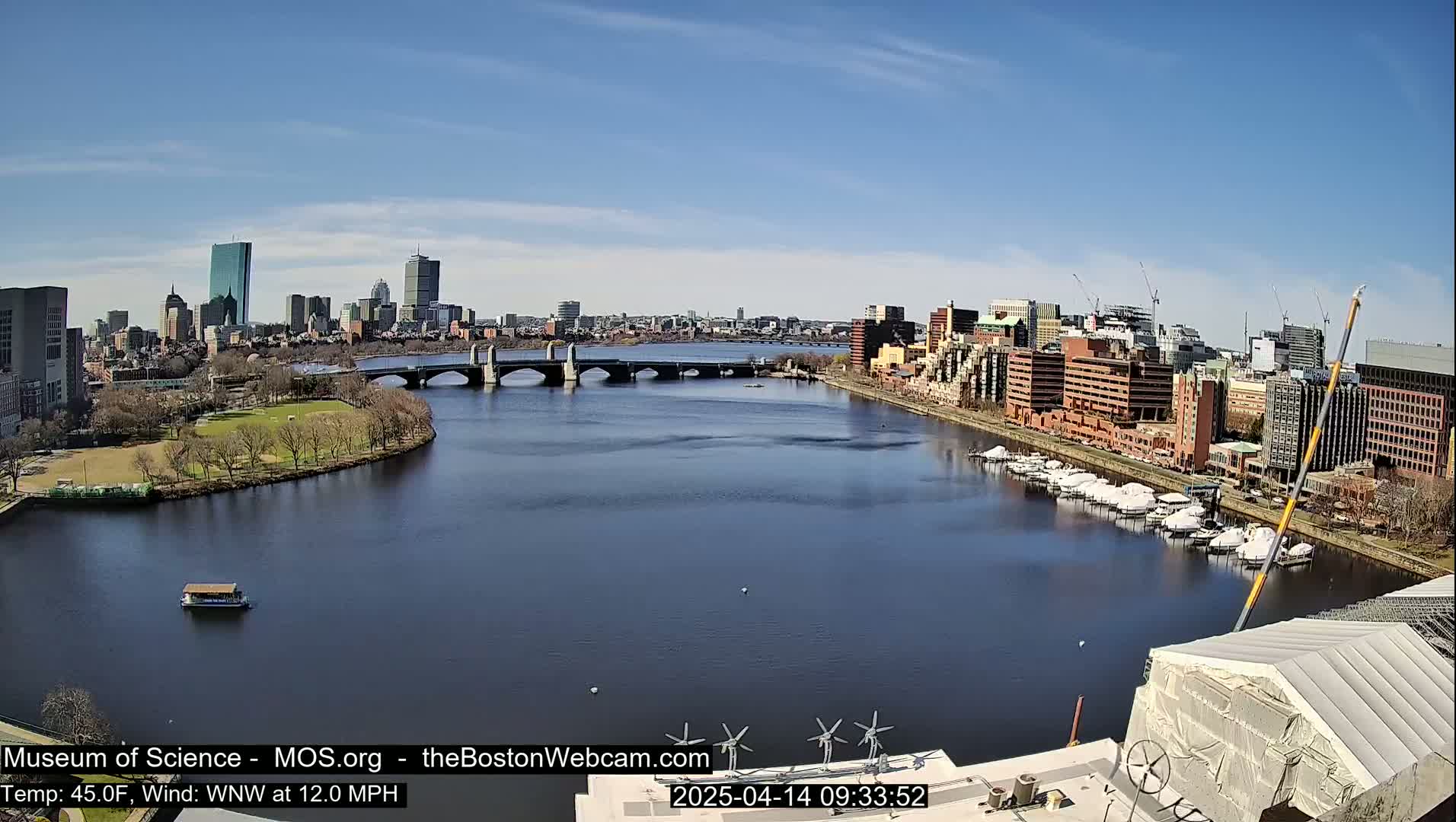A sunny cityscape with a river, bridge, and boats, under a clear blue sky.