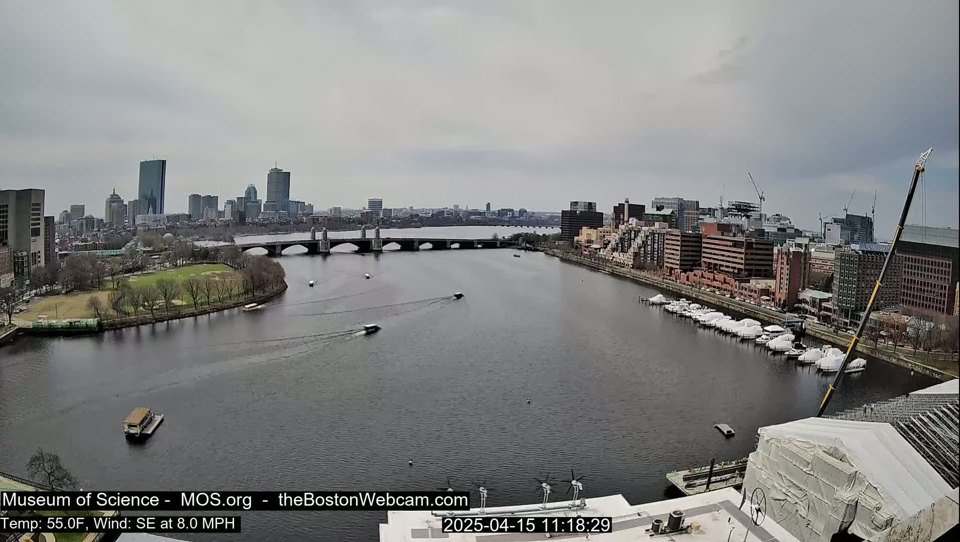 A wide shot shows a river with boats and a bridge leading to a city skyline under an overcast sky.