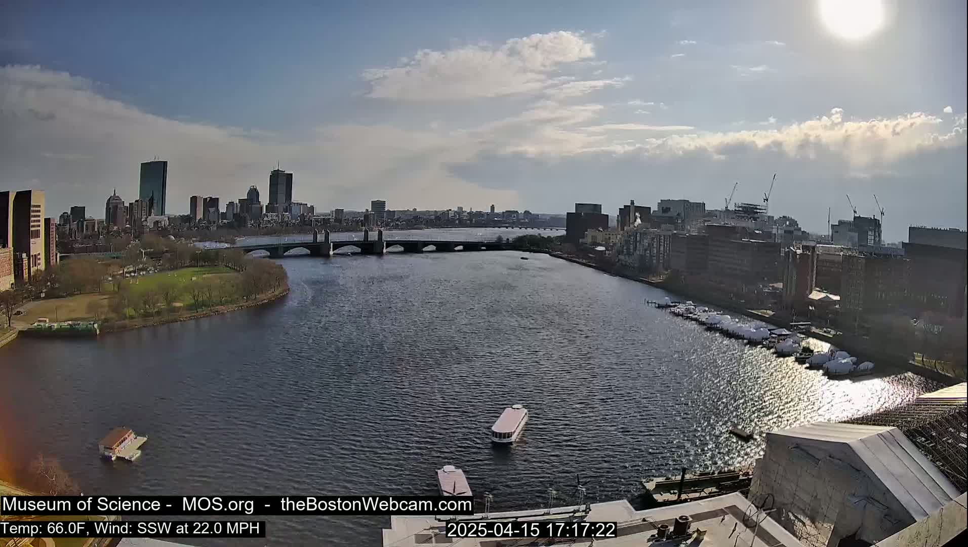 A sunny day shows a city skyline across a wide river with a bridge, boats docked, and a few small vessels on the water.